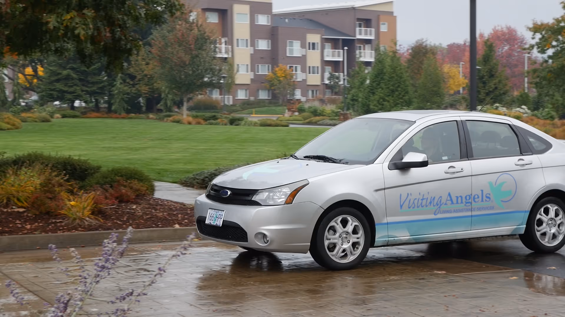 Silver Visiting Angels car parked on a wet driveway near a green lawn and apartment buildings