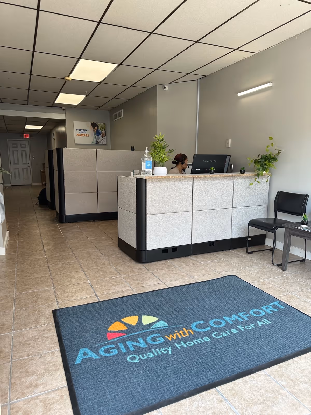 Reception area with a desk, staff at computer, seating, and Aging With Comfort mat.