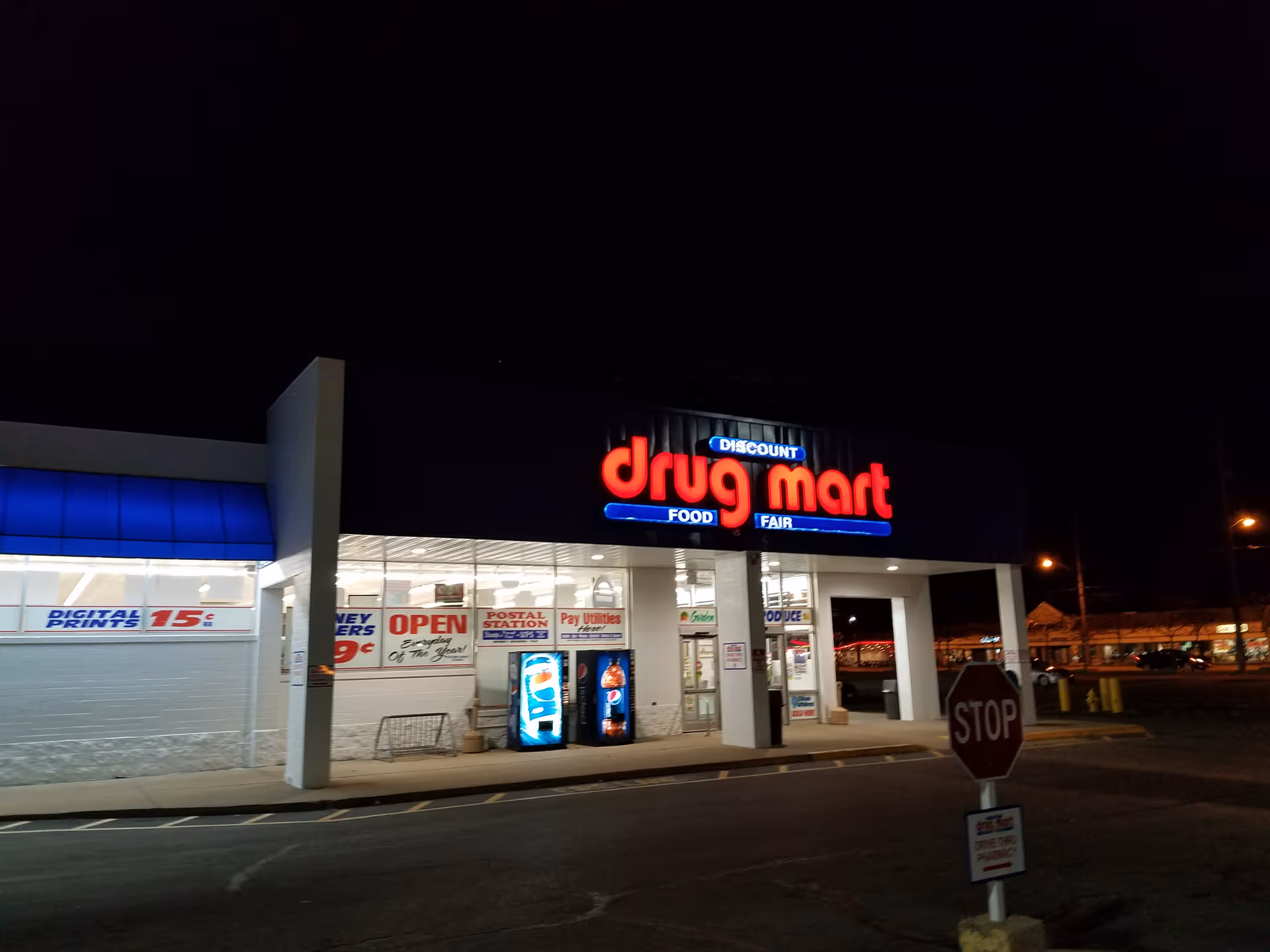 Night exterior of Discount Drug Mart storefront with illuminated red sign and blue awning.