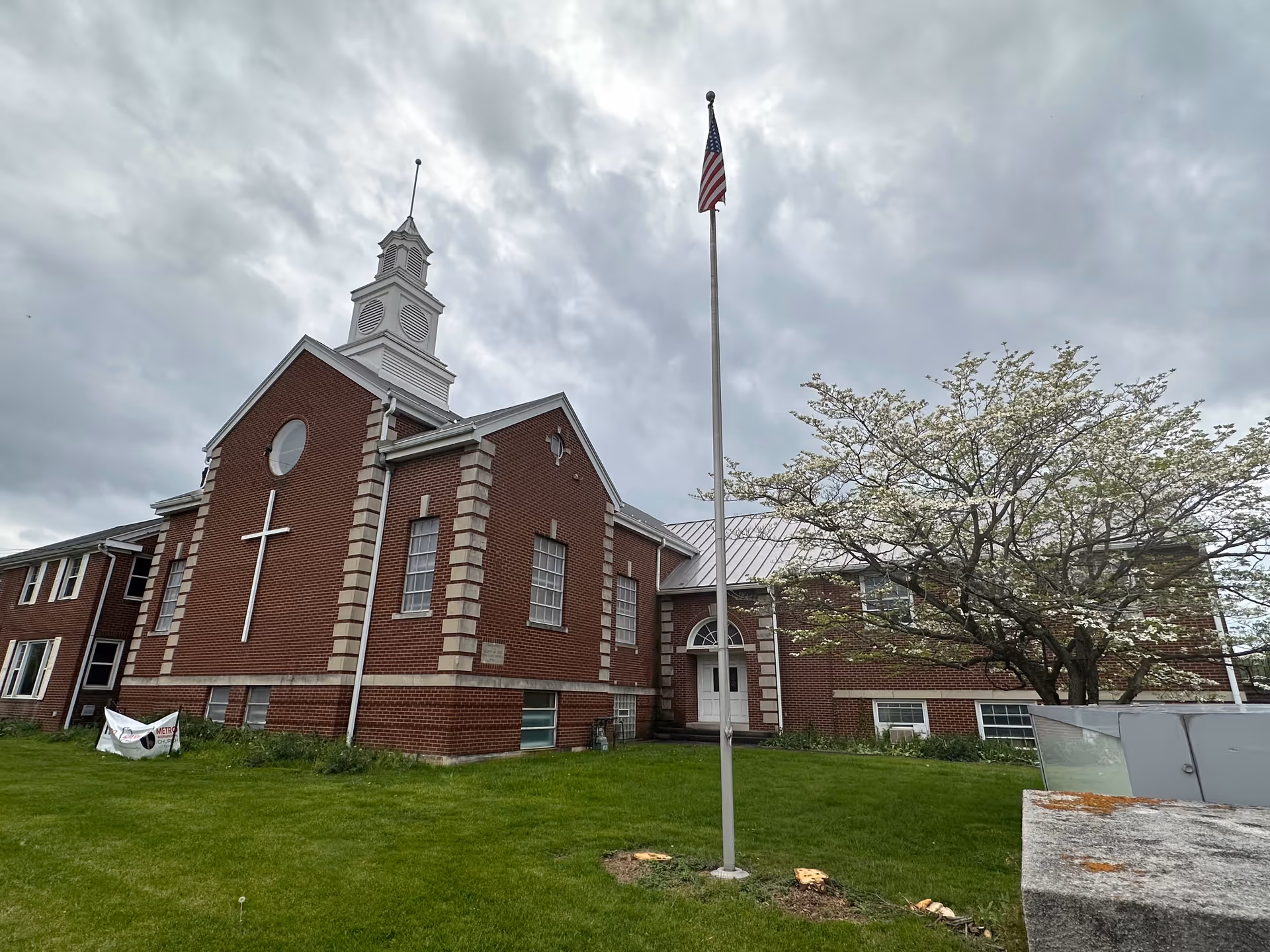 Brick building with a cross on the front, flagpole, and blossoming tree under cloudy sky.