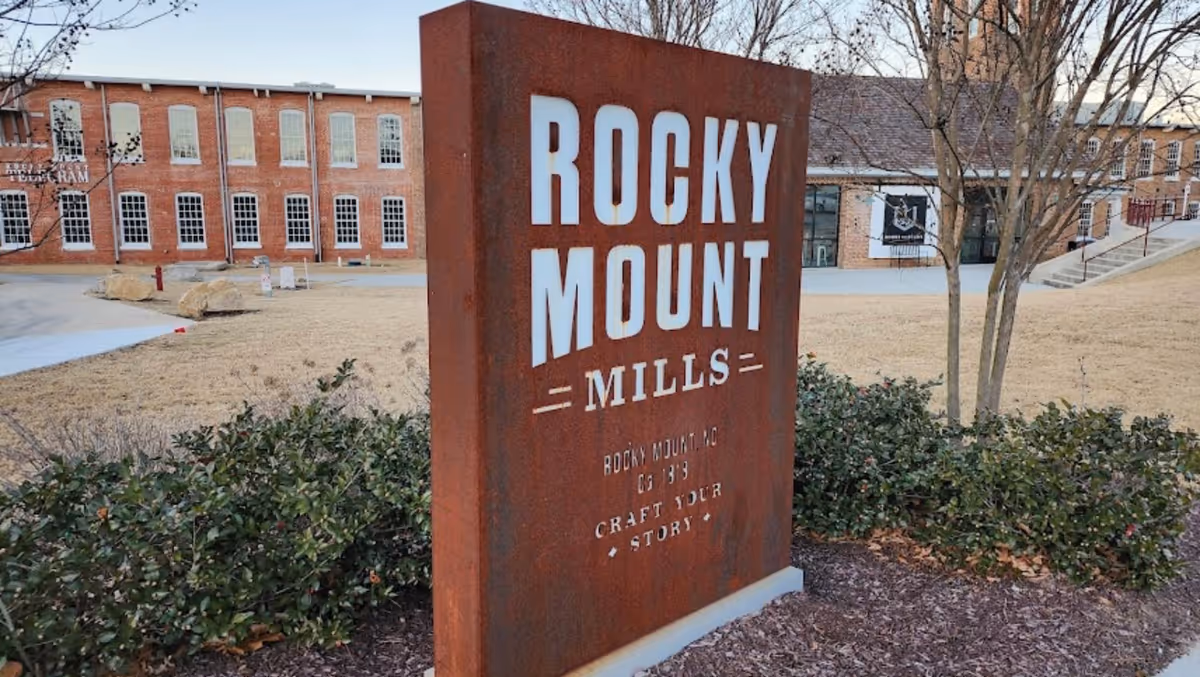A large rust-colored Rocky Mount Mills sign outdoors with brick buildings in background.