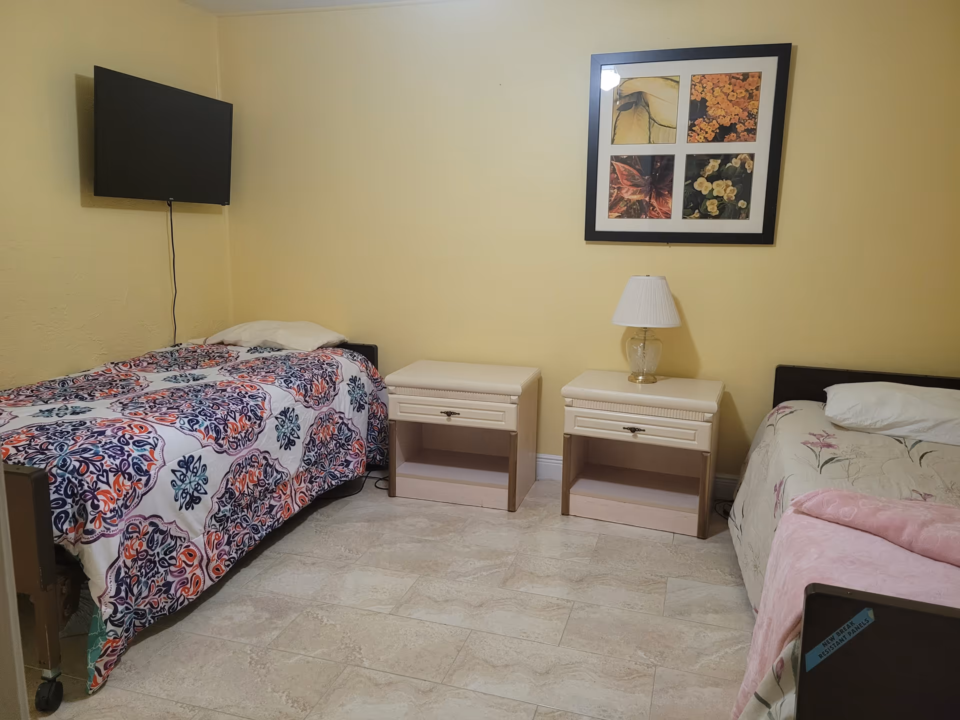 A two-bed senior living bedroom with wall-mounted TV, nightstands, lamp, and framed art.