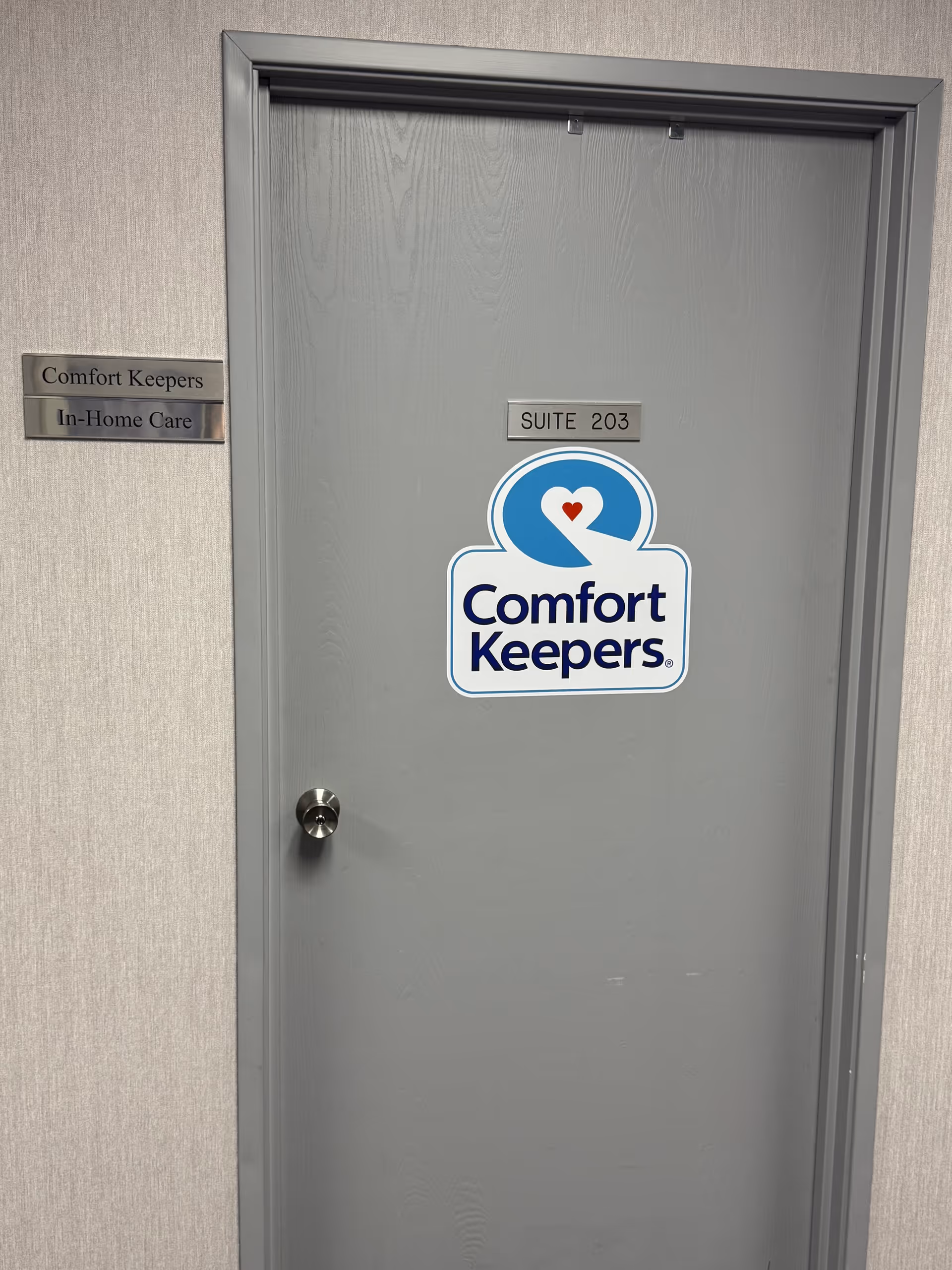 Gray door with Comfort Keepers sign and suite number in an interior hallway.