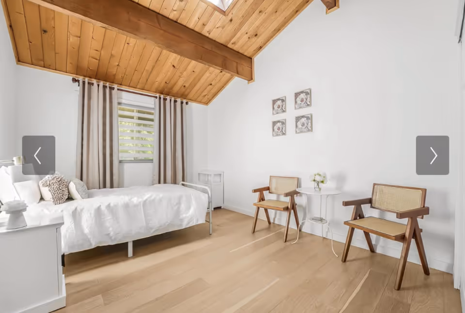 Bright bedroom with white linens, wooden beams, and two chairs by a small table.