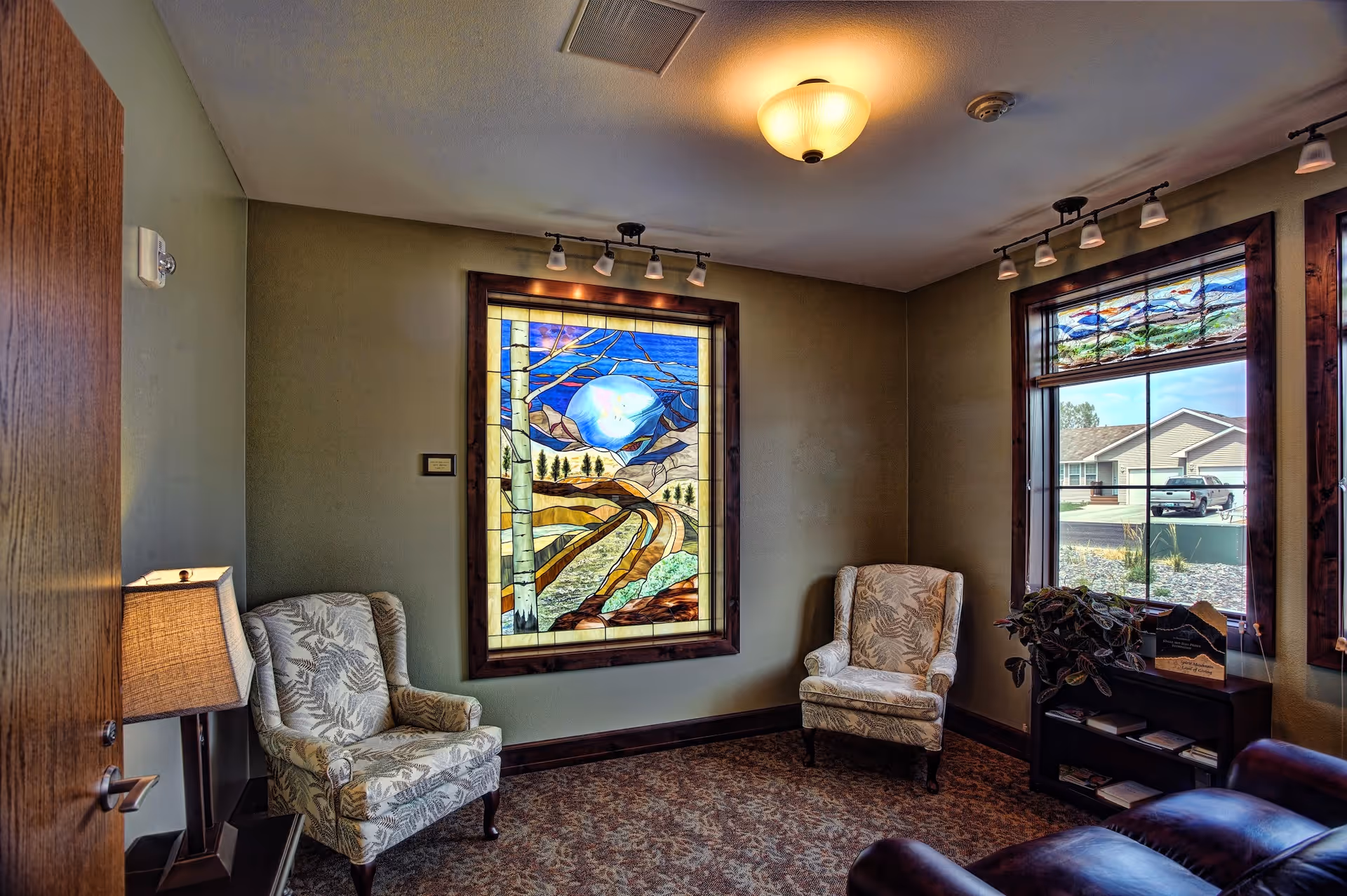 Cozy lounge area with patterned chairs, stained glass window, and magazines by a lamp.