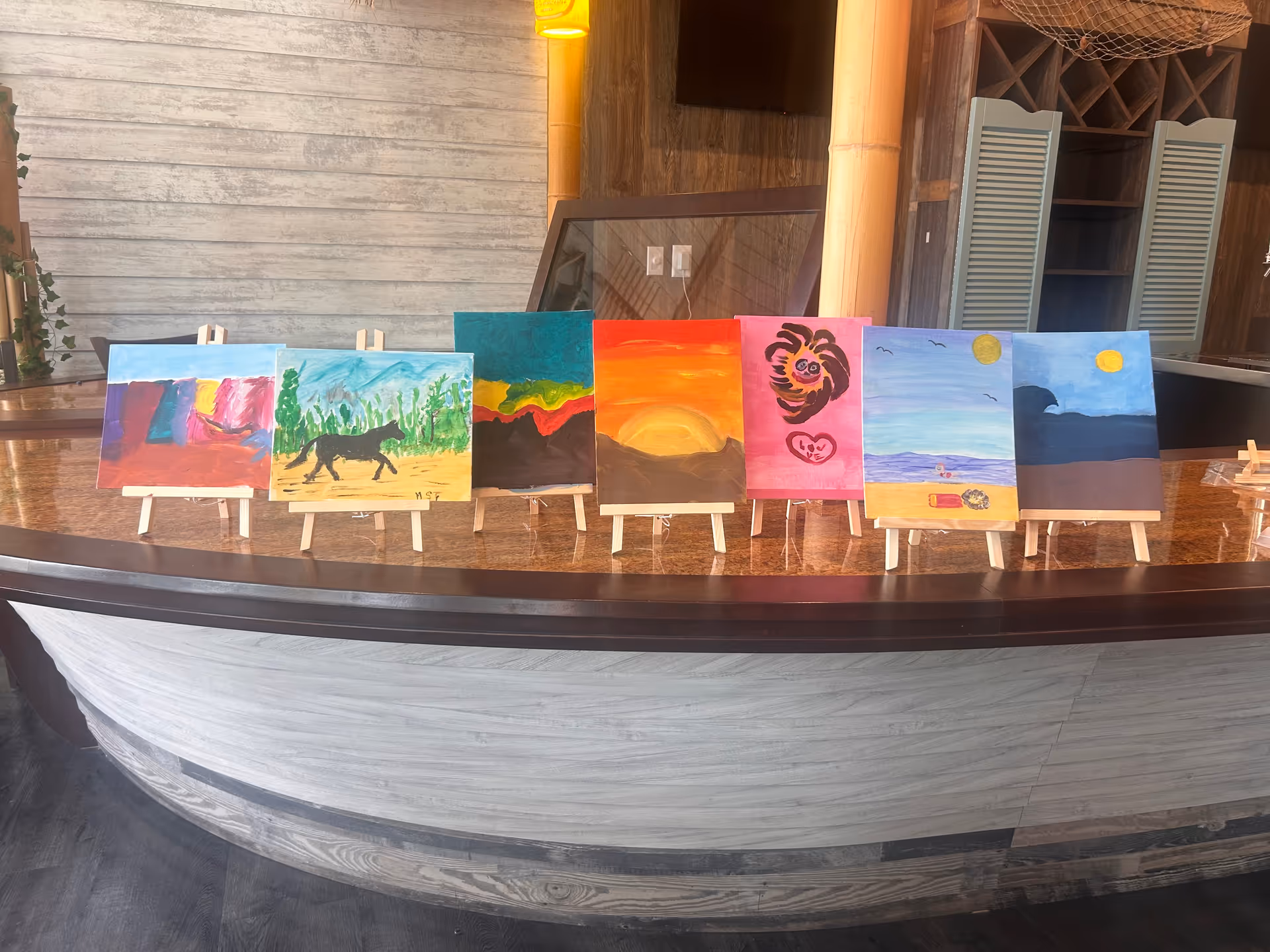 Colorful paintings on small easels displayed along a curved counter inside a facility.
