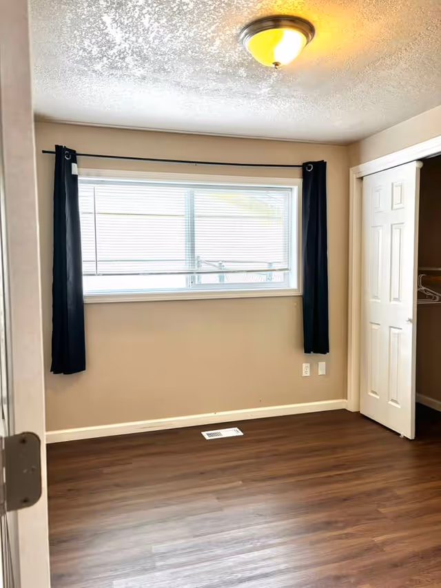 An empty bedroom with dark curtains, a window, wood flooring, and a closet.