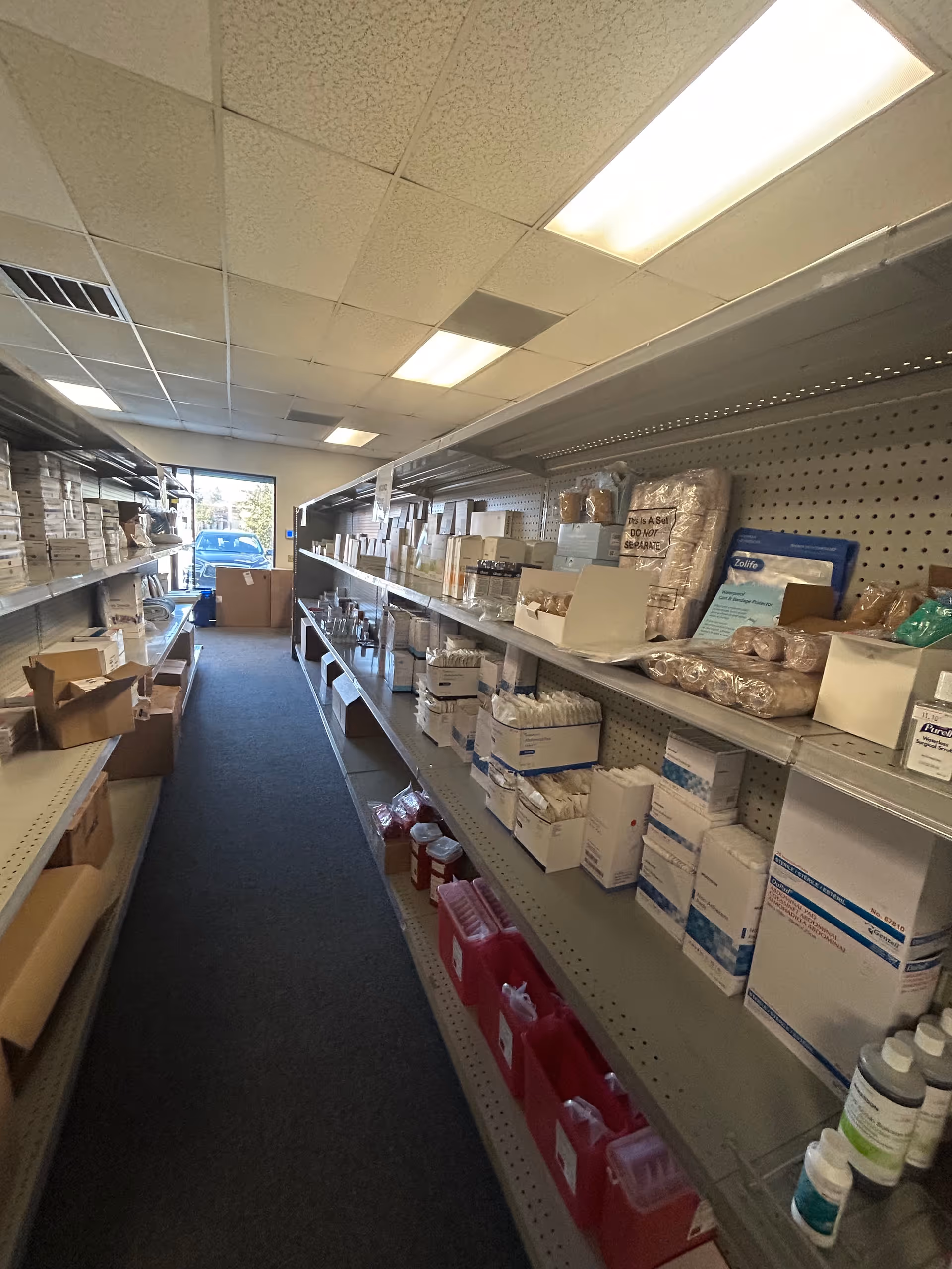 Narrow aisle with metal shelves stocked with medical supplies in a storage room.
