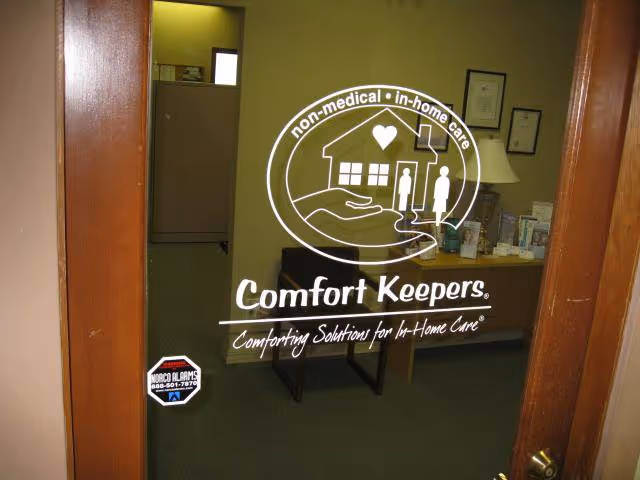 Indoor reception area seen through a glass door displaying the Comfort Keepers logo.