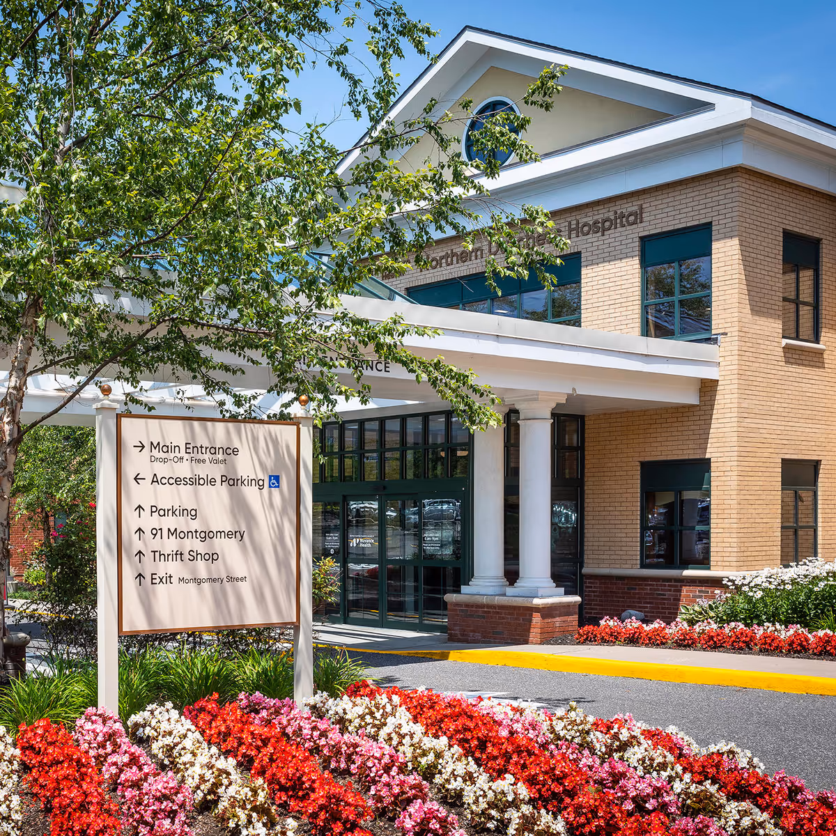 Exterior hospital entrance with glass doors, signage, columns, and vibrant flower beds.