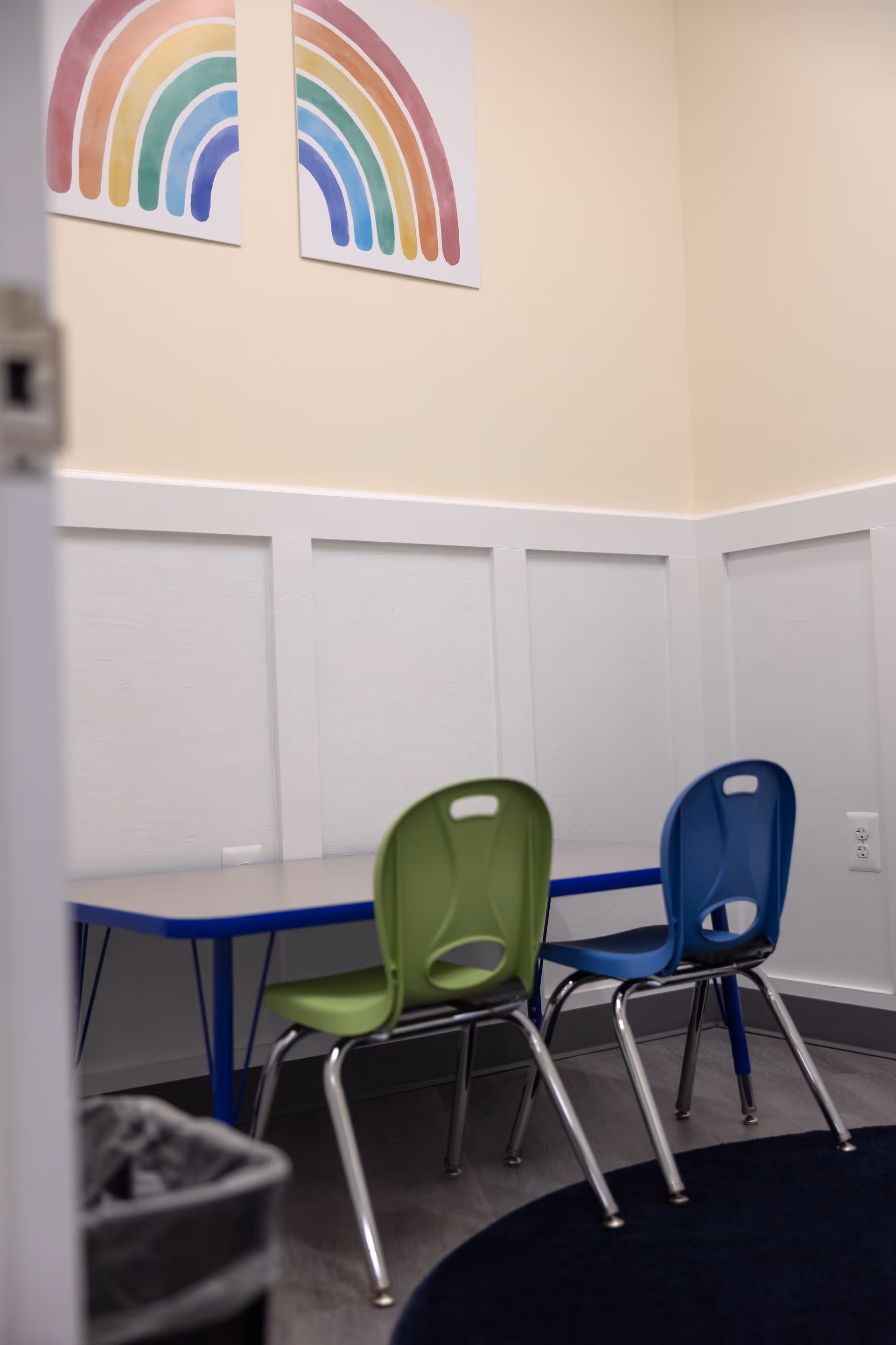Two colorful chairs sit beside a blue table in a beige-walled room.