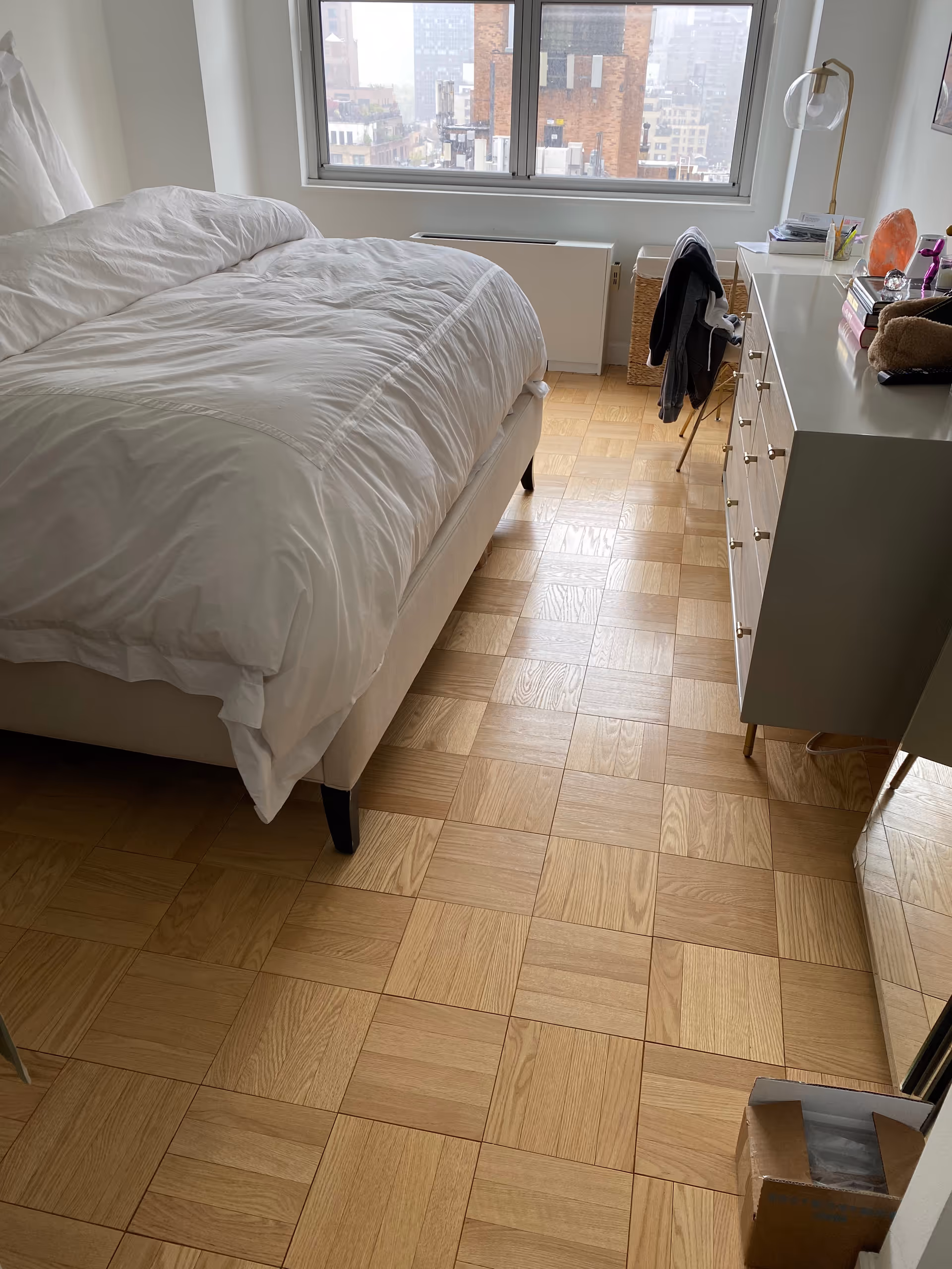 A neatly made bedroom with a bed, dresser, and large window overlooking a city skyline.