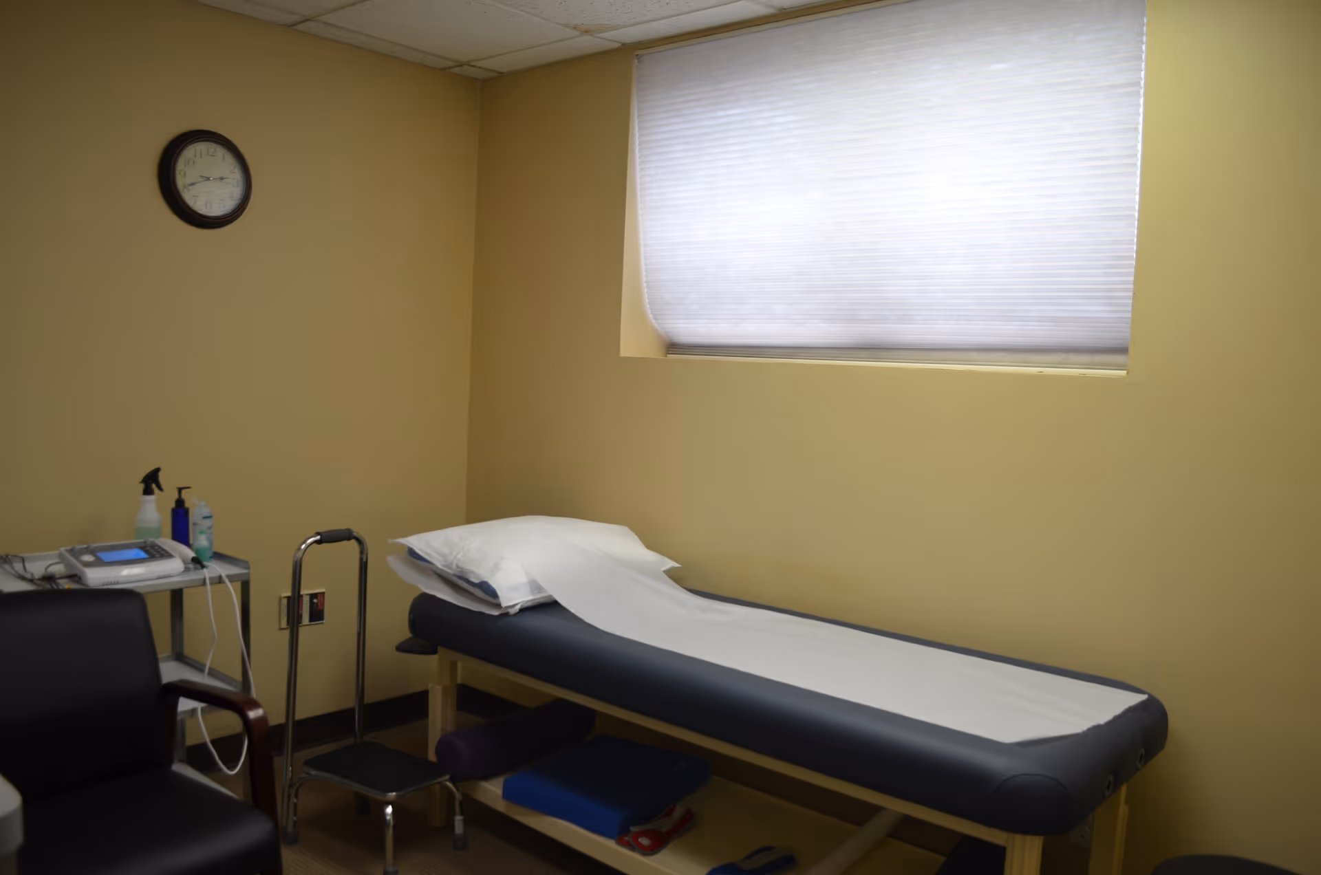 A small therapy room with an exam bed, cart, clock, and window shade.