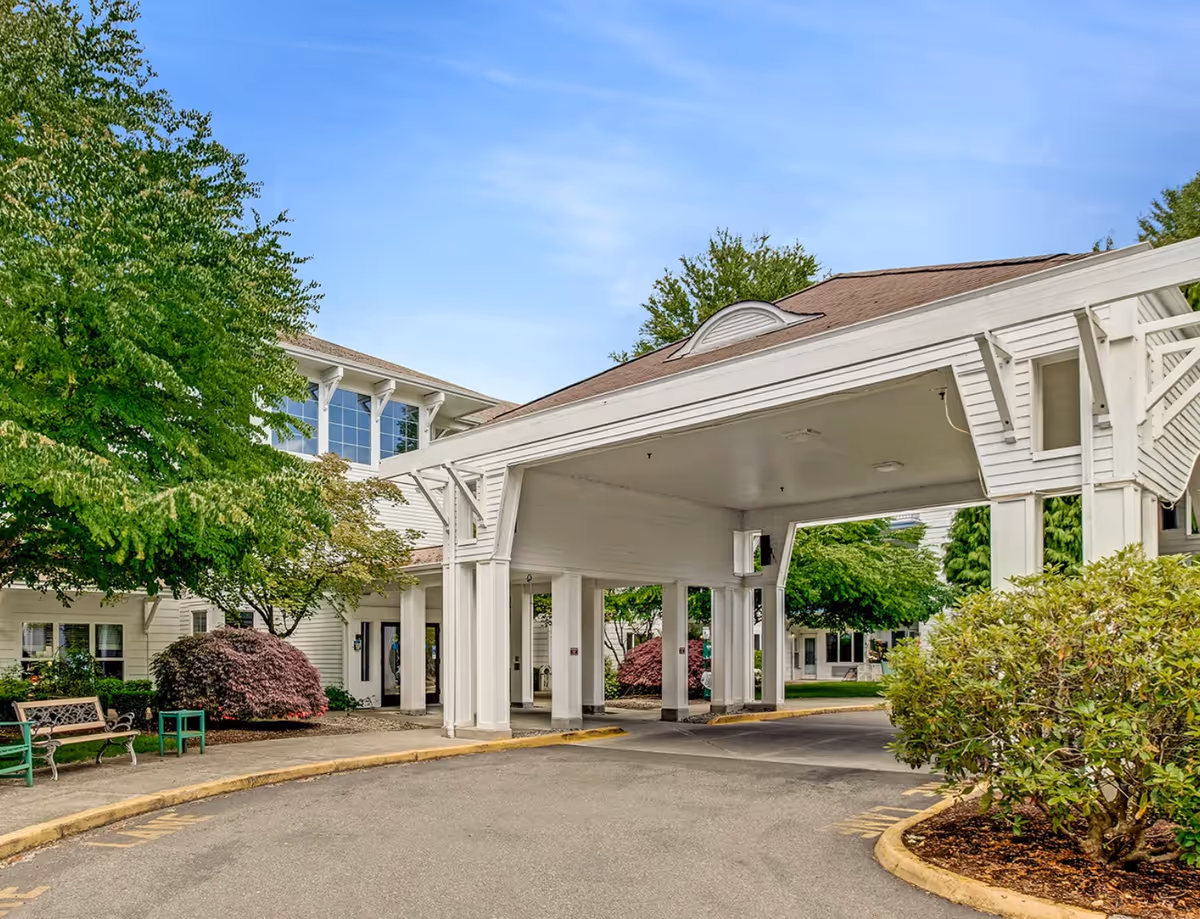 White building with a covered entrance, driveway, and lush shrubs.