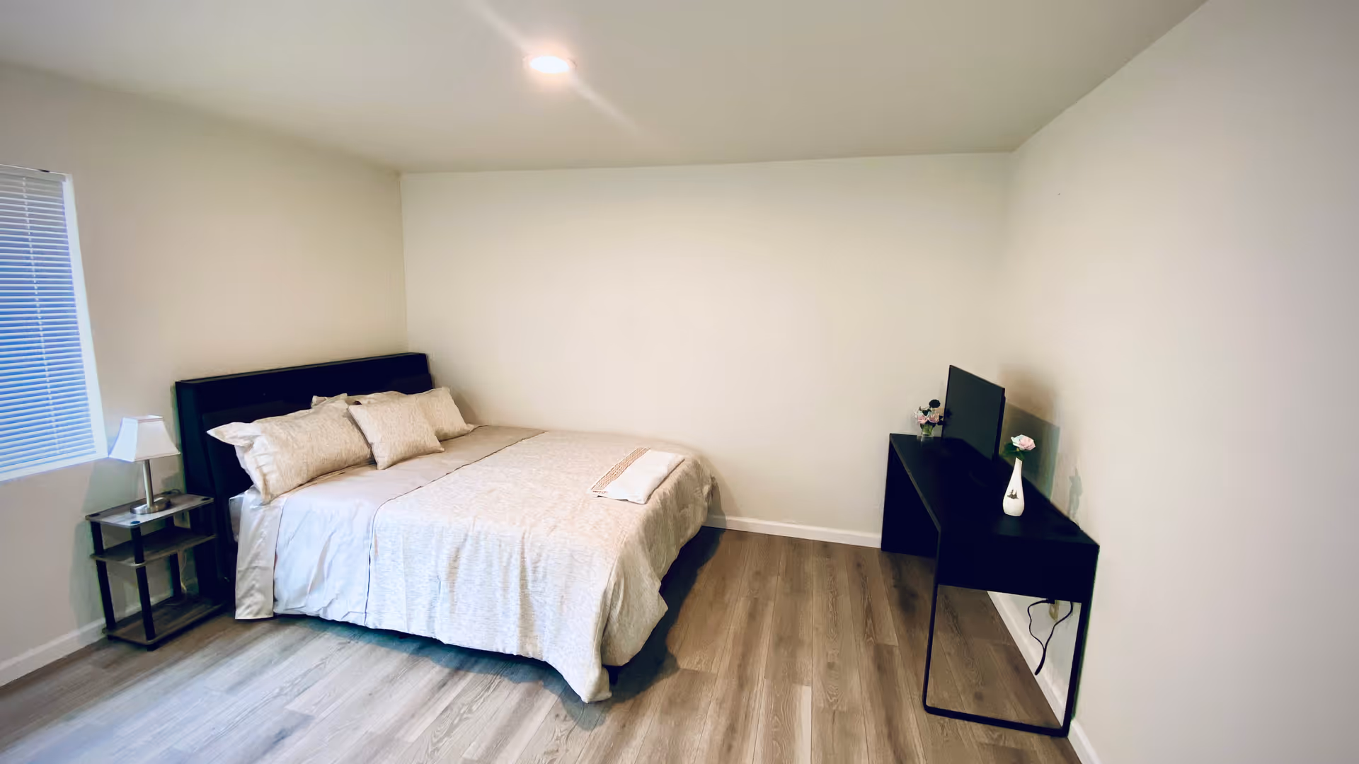 A small, minimalist bedroom featuring a bed, nightstand, desk, and window in a clean, neutral space.