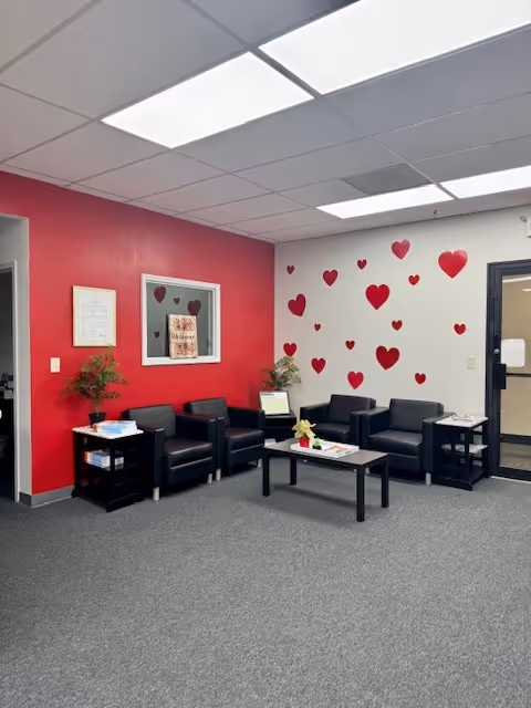 Nursing facility waiting lounge with red wall, heart decals, black chairs, tables, and plants.