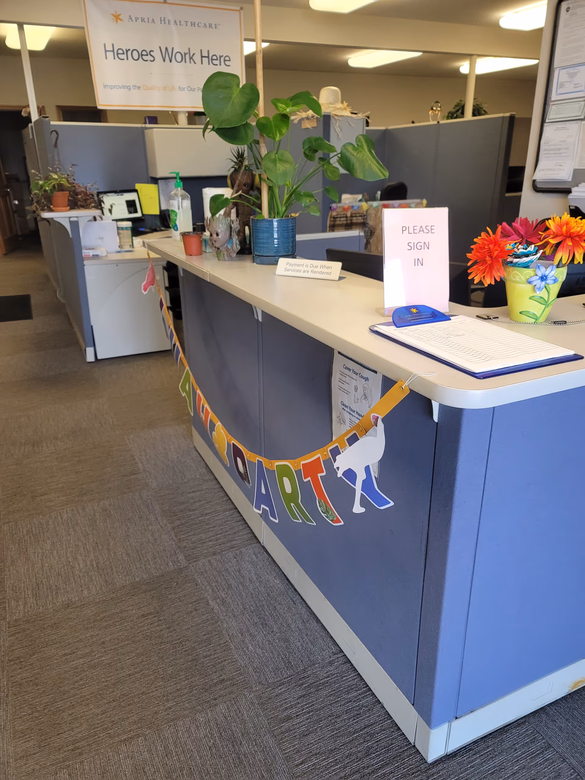 Reception desk in a healthcare facility with signage, plants, and sign-in materials.
