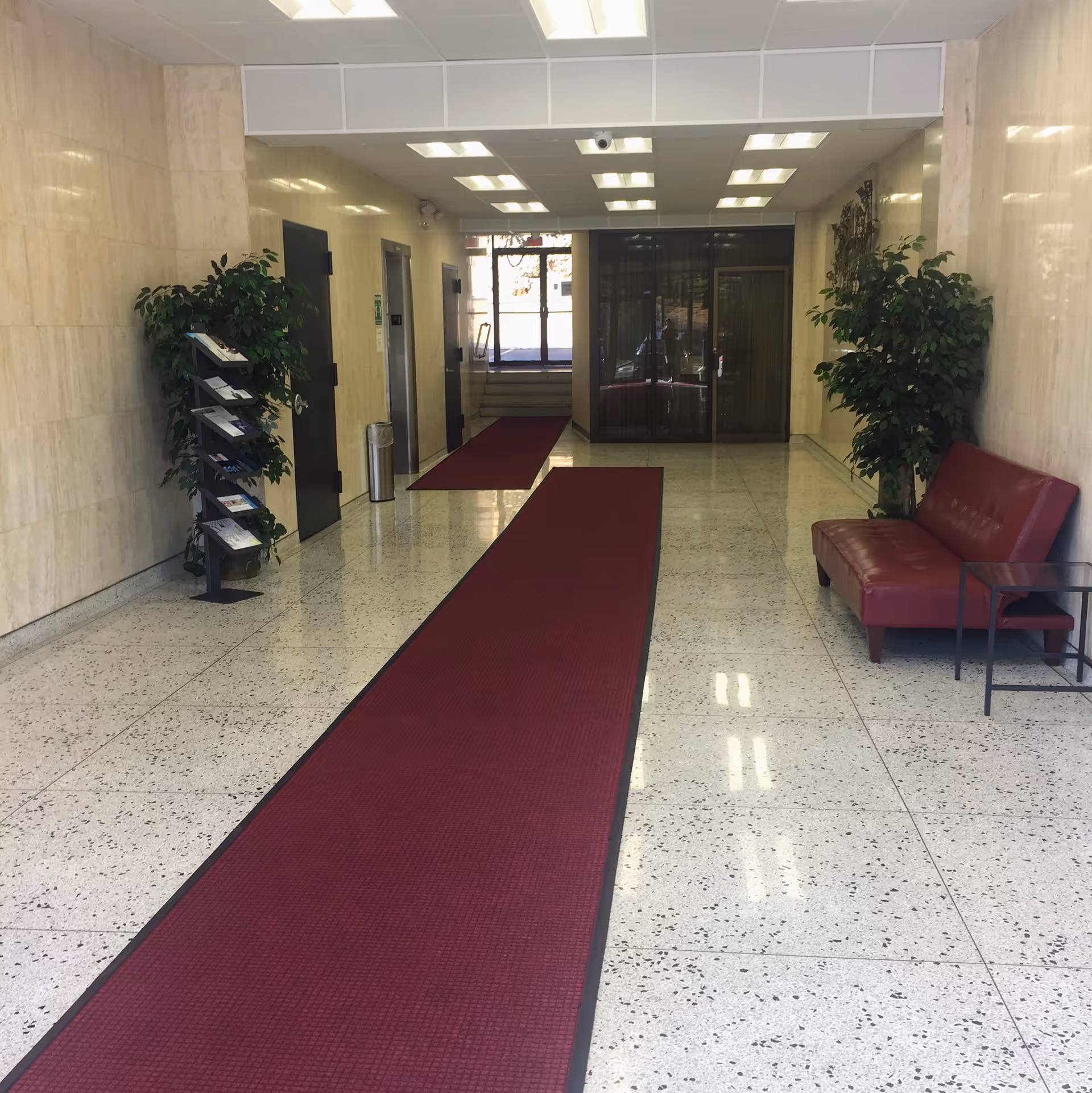 Bright marble hallway with a red carpet runner, plants, and red seating near glass doors.