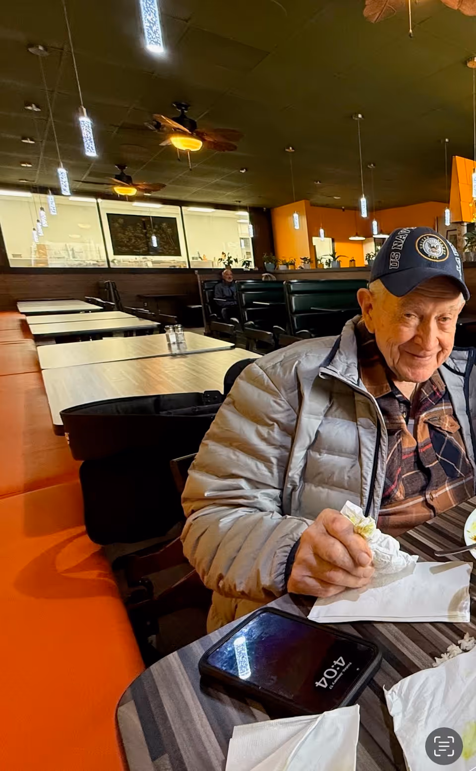 Elderly man sits at a table inside a busy dining room of a senior facility.