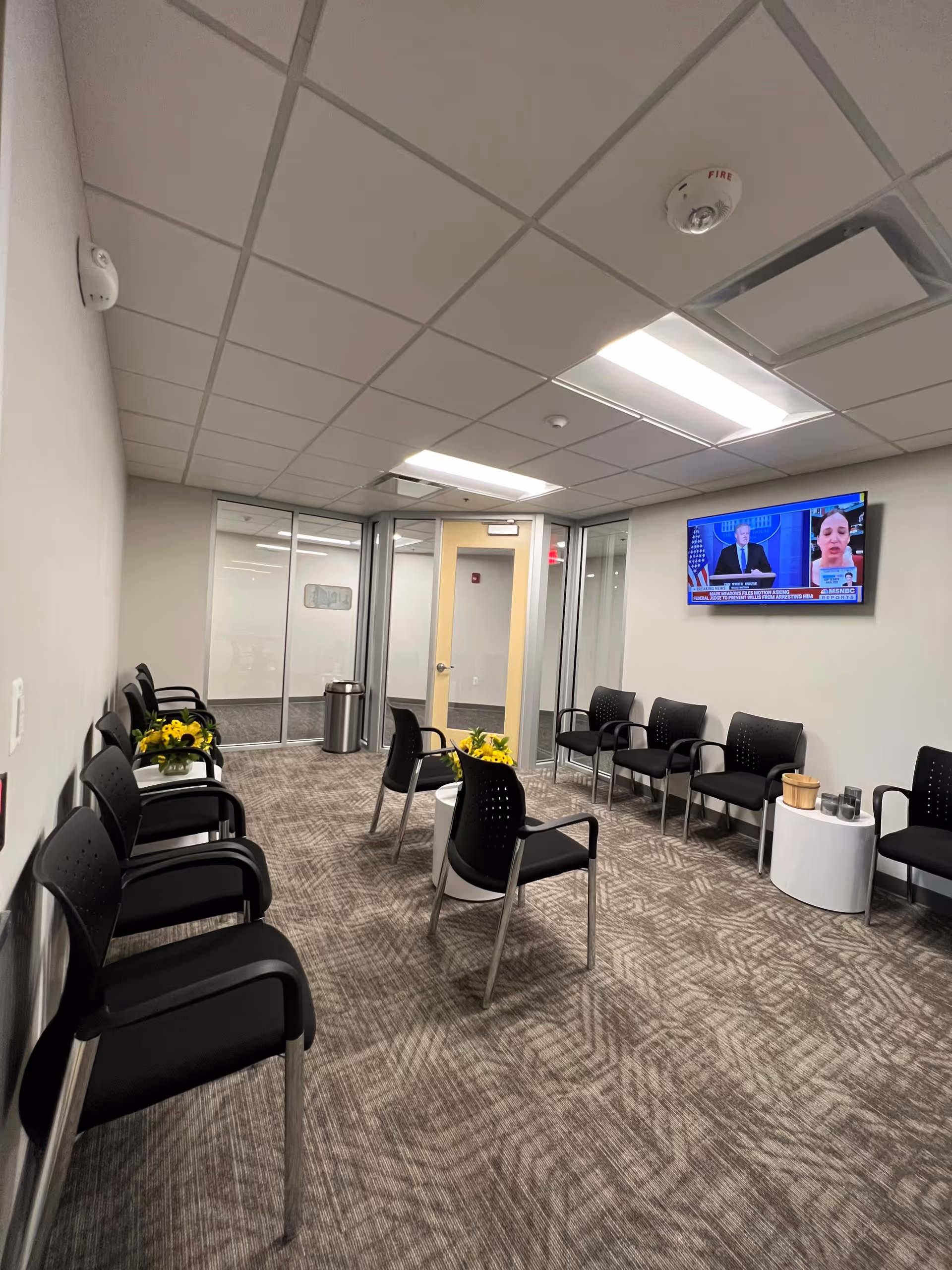 Interior waiting area with black chairs, a wall TV, glass doors, and yellow flower arrangements.