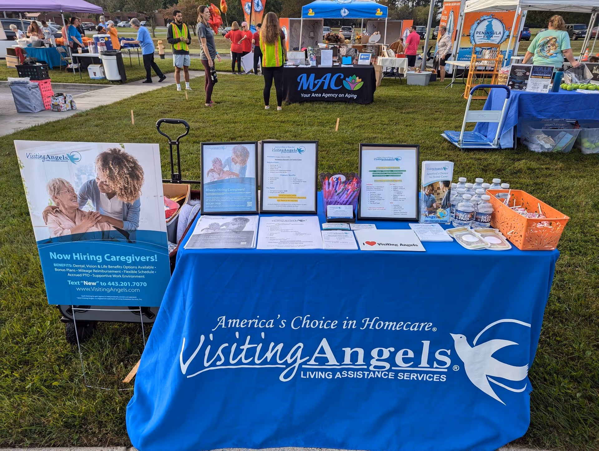 Outdoor vendor booth for Visiting Angels Senior Home Care with banners, tables, and people milling about.