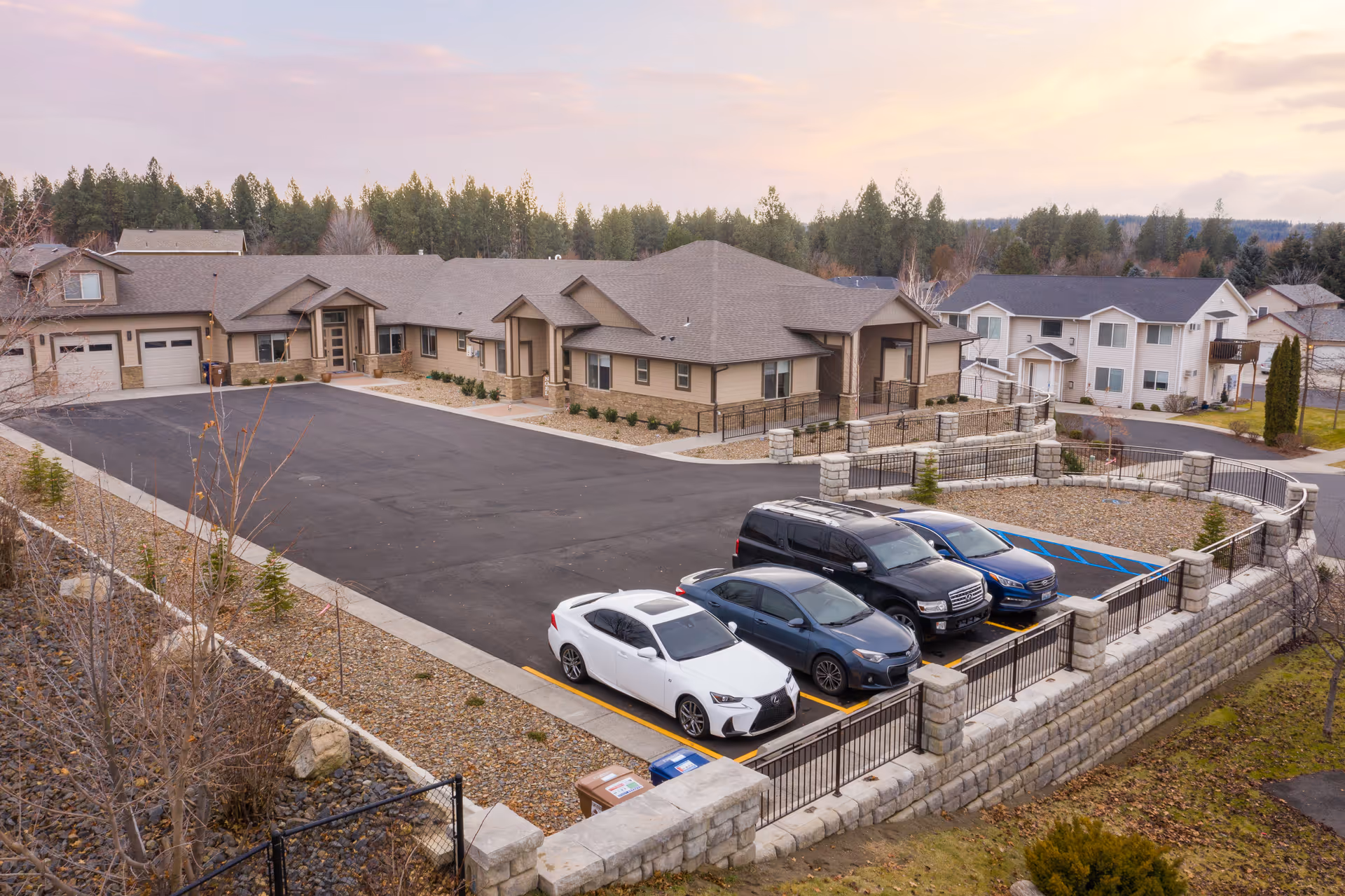 A residential-style senior living campus with beige buildings, a paved parking lot, and parked cars.
