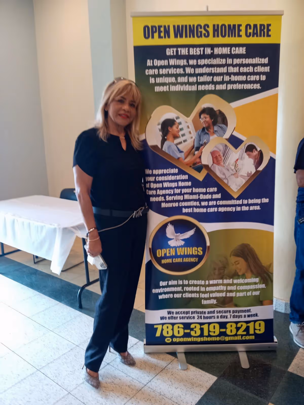 A woman stands beside a tall Open Wings Home Care banner inside a facility corridor.