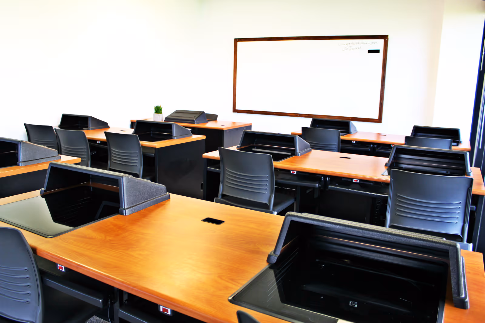 Empty classroom with wooden desks and black chairs arranged in rows, whiteboard on wall.