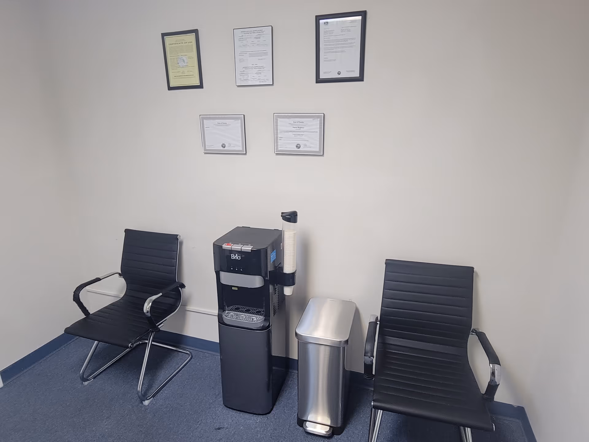 Two black chairs surround a coffee machine and trash bin against framed certificates on the wall.