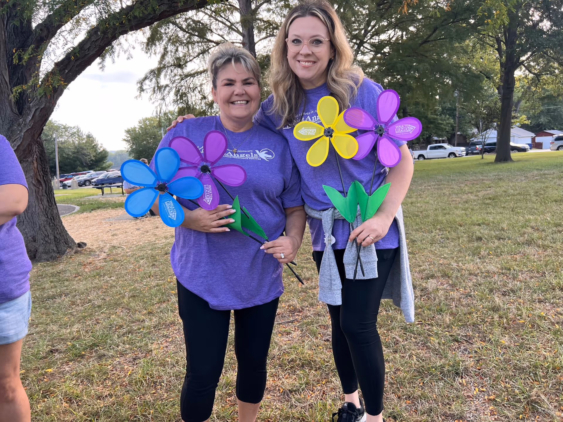 Two smiling caregivers hold colorful pinwheels outdoors in a park-like setting.