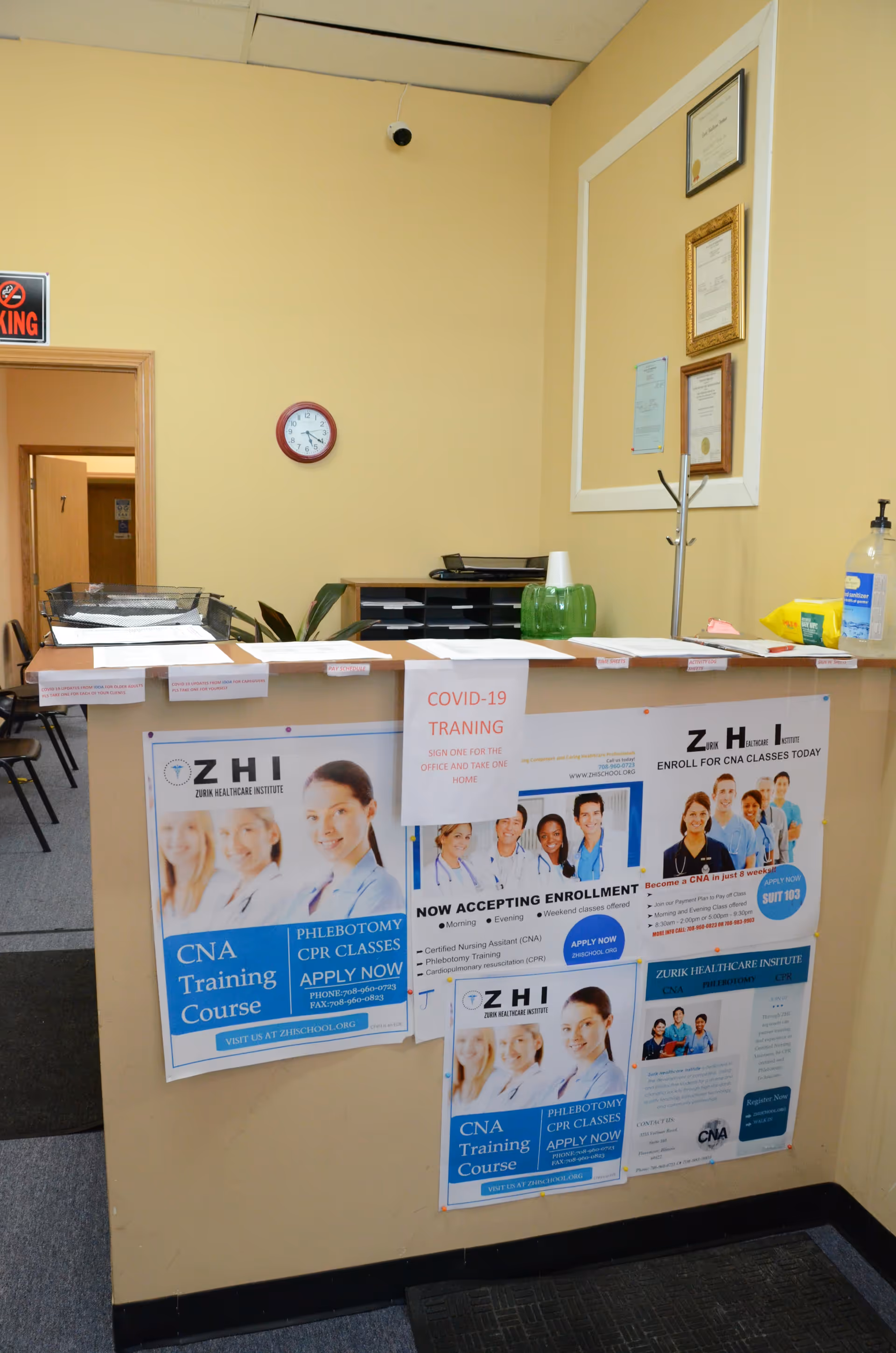 Reception desk with posters advertising CNA training in a yellow-walled healthcare facility lobby.