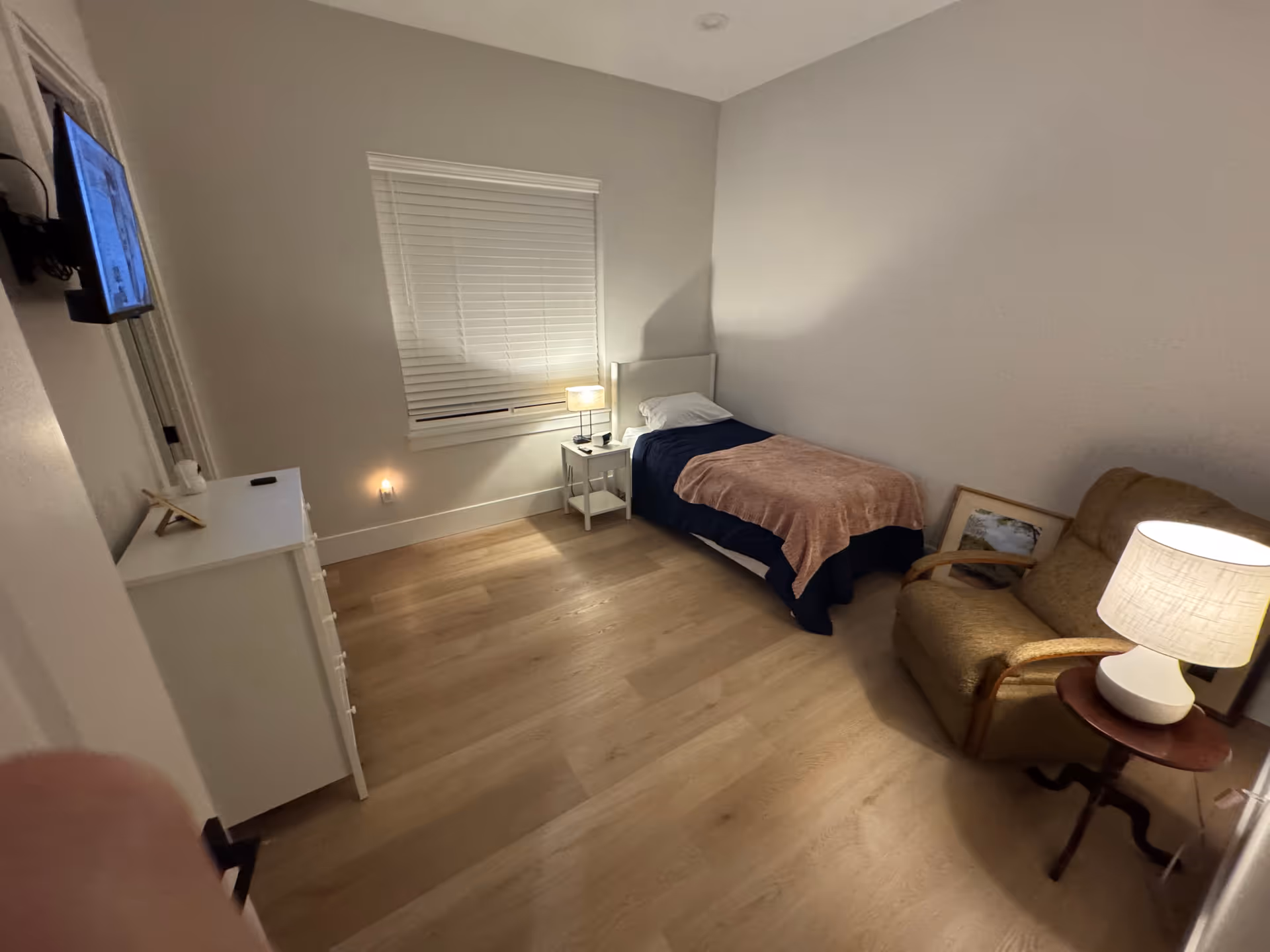 Compact bedroom with a single bed, nightstand, dresser, and seating.