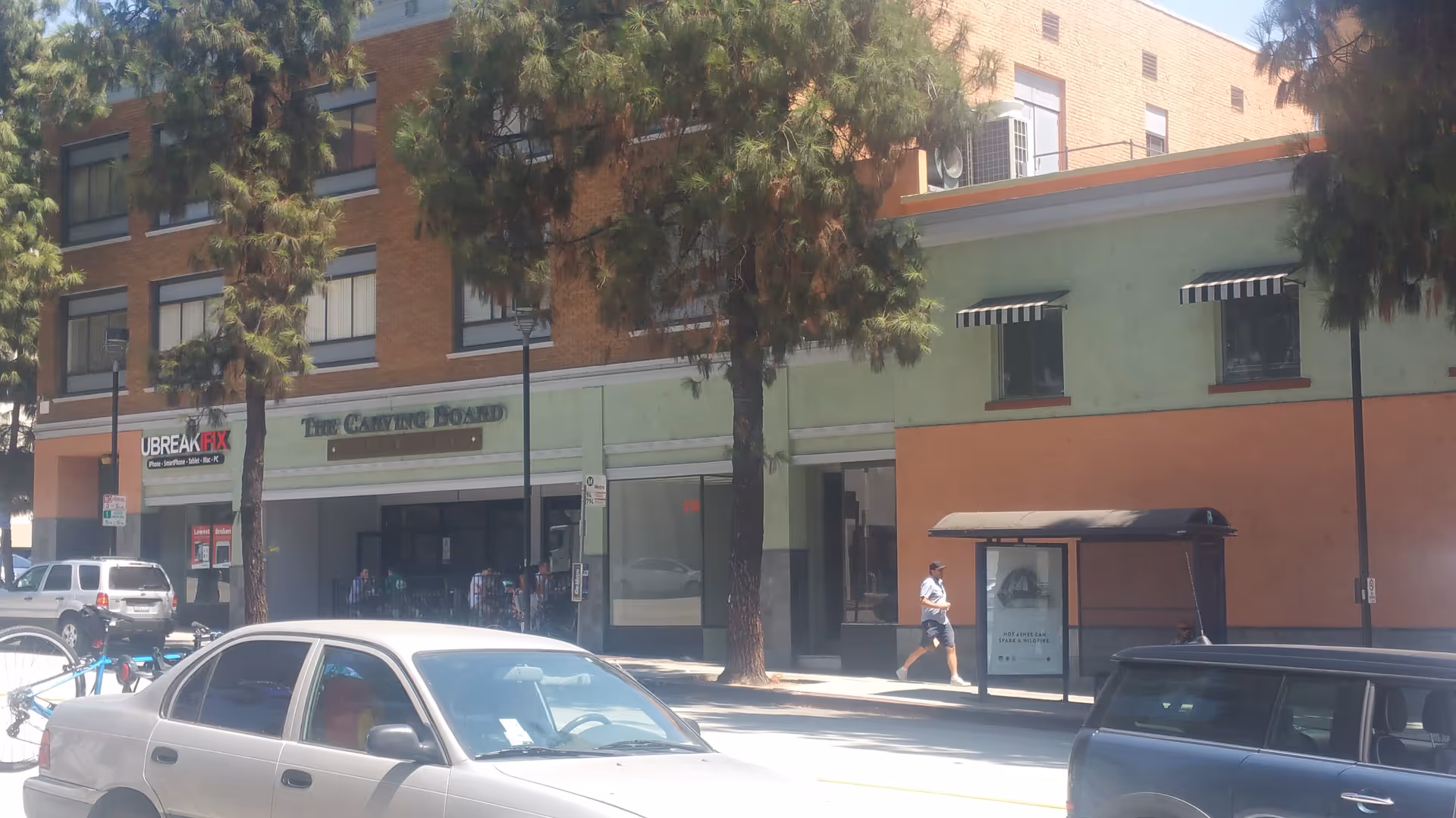 Exterior street view showing storefronts, parked cars, trees, and a bus stop along the sidewalk.