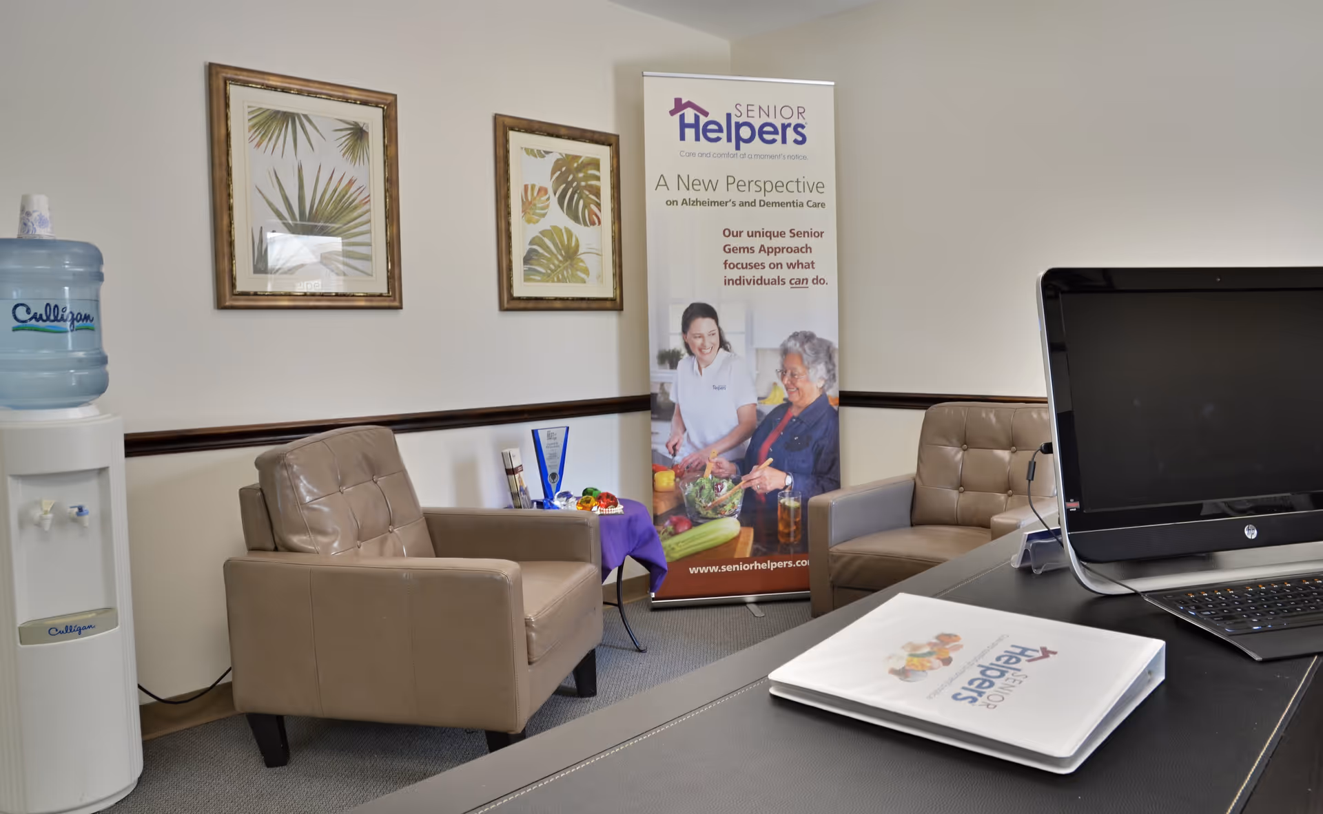 Reception area with beige chairs, water cooler, banner, and a laptop on a desk.