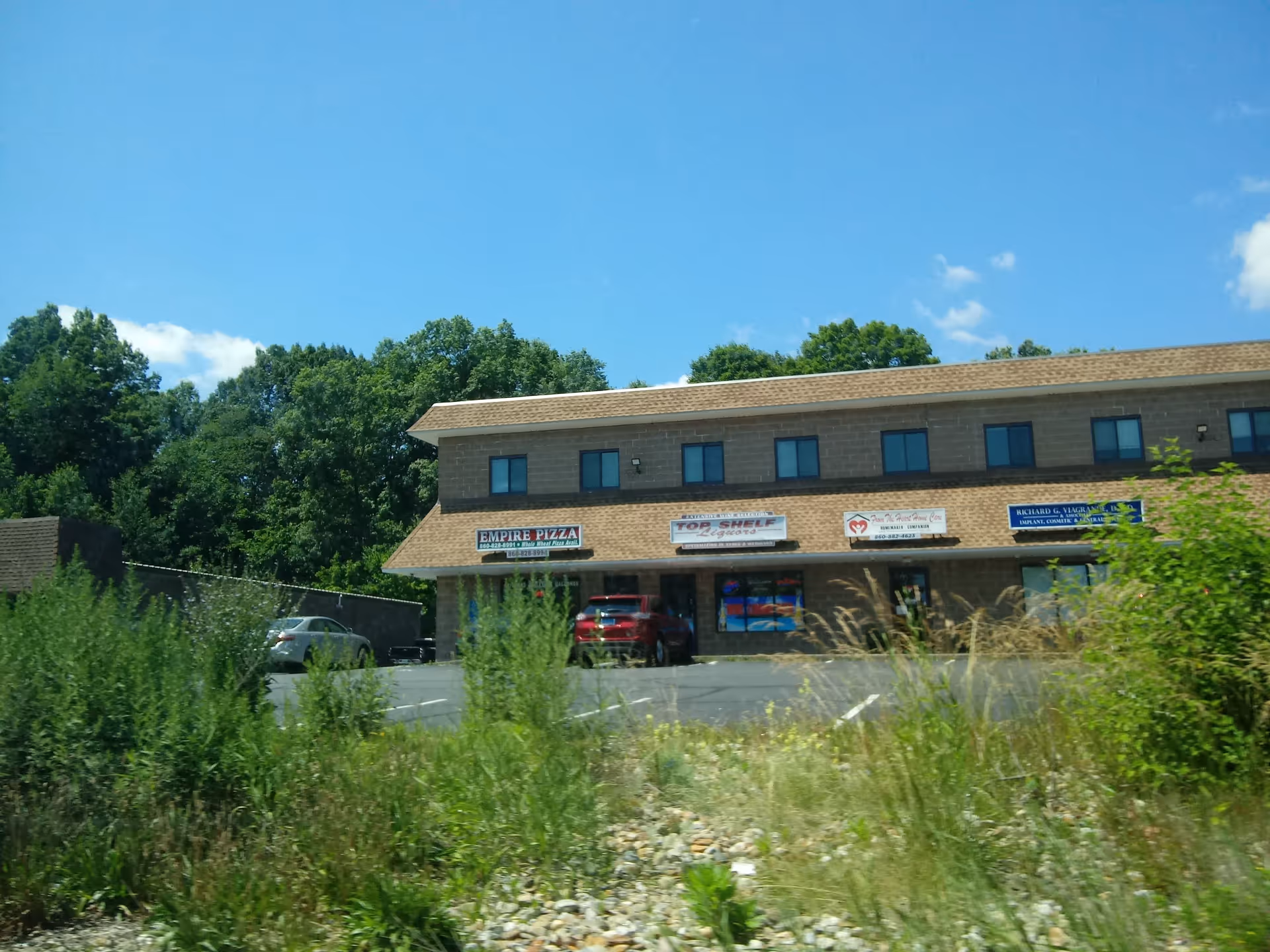 Two-story commercial building with signs, parked cars, and overgrown foreground vegetation under blue sky