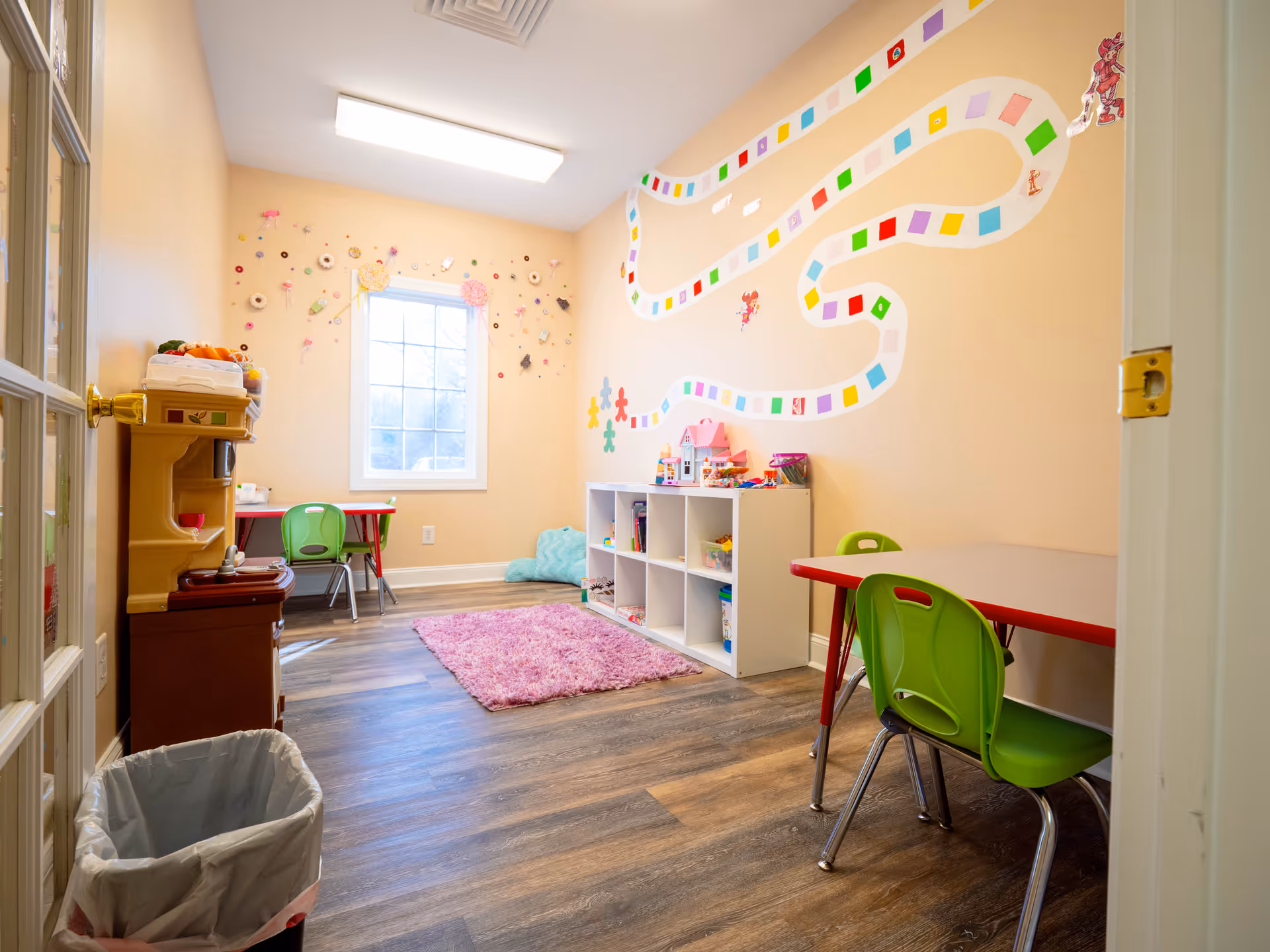 Bright children's playroom with colorful wall mural, tables, chairs, and shelves.