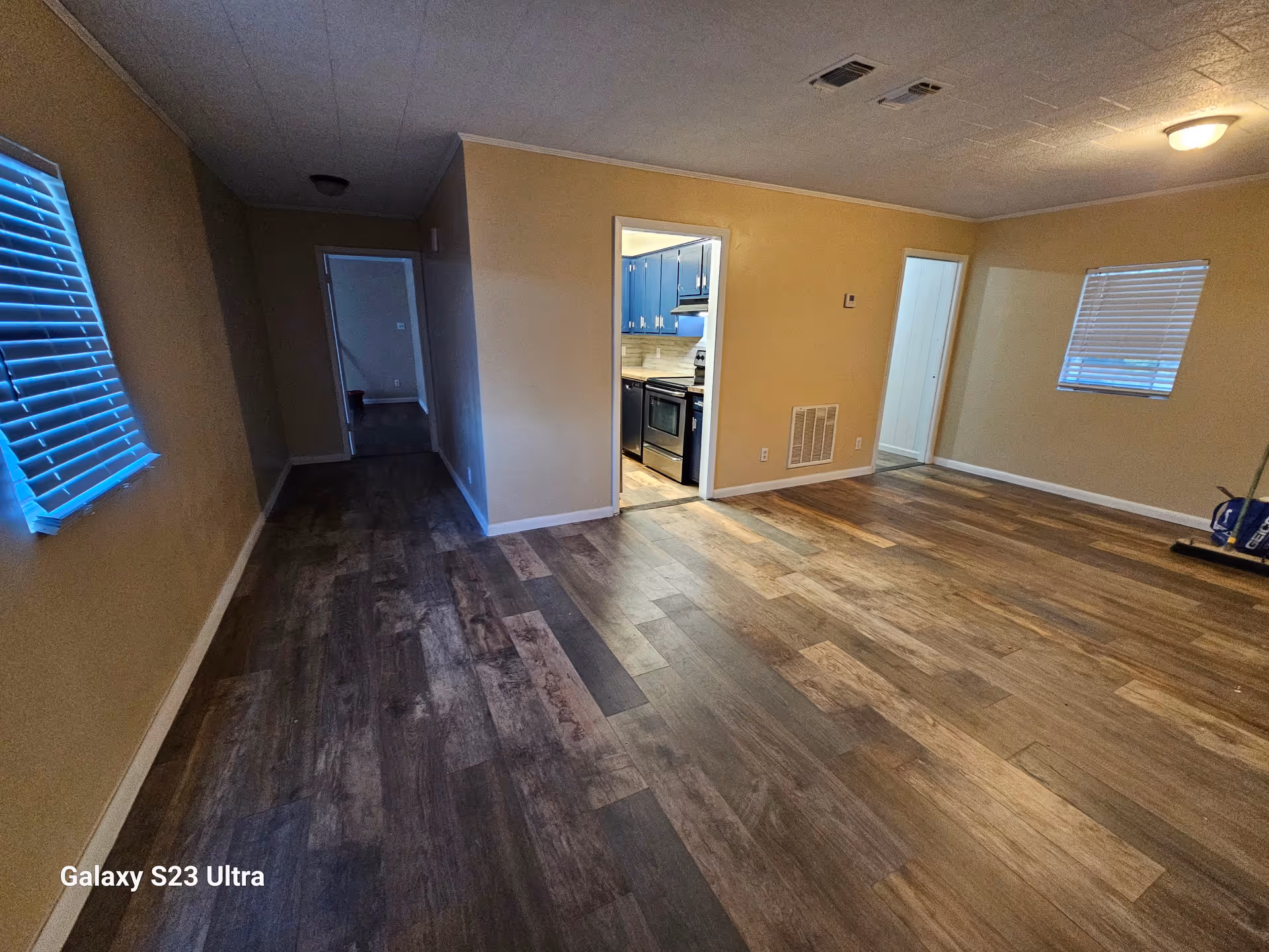 Spacious living area with wood laminate floors, beige walls, and a visible kitchen doorway.
