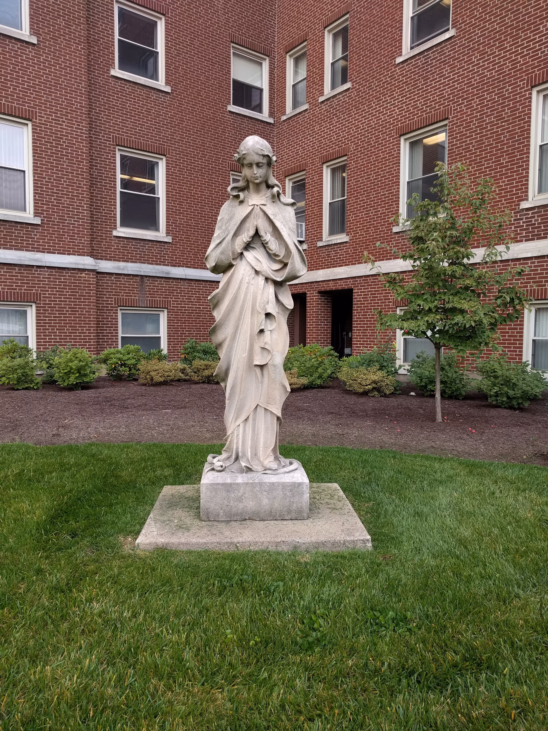 Stone statue of a robed figure stands on a pedestal in a grassy courtyard.
