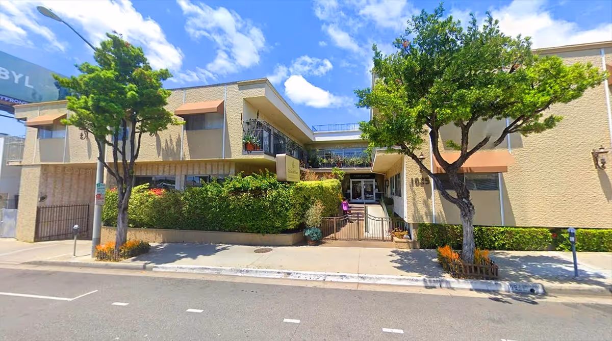 Two-story beige senior living building with glass entrance, trees, and shrubbery along the sidewalk.