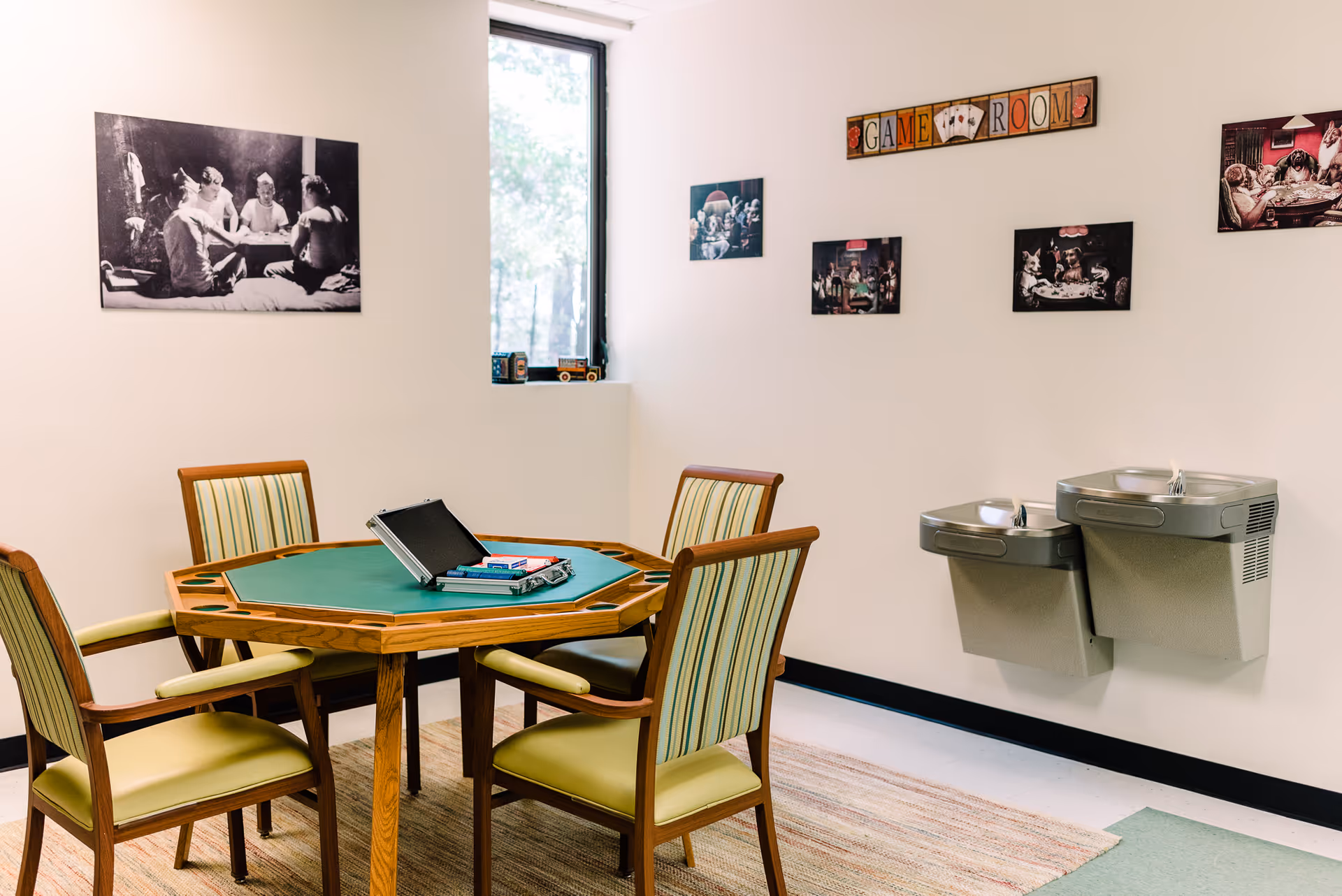 Indoor game room with round green-felt table, four cushioned chairs, wall art, and drinking fountains.