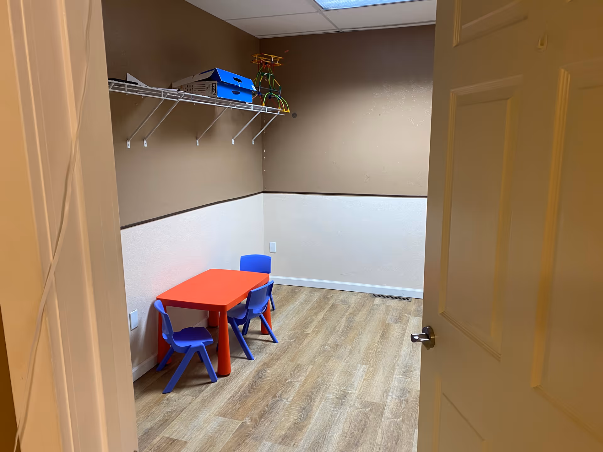 Small activity room with a red table and blue chairs, toys on shelf.