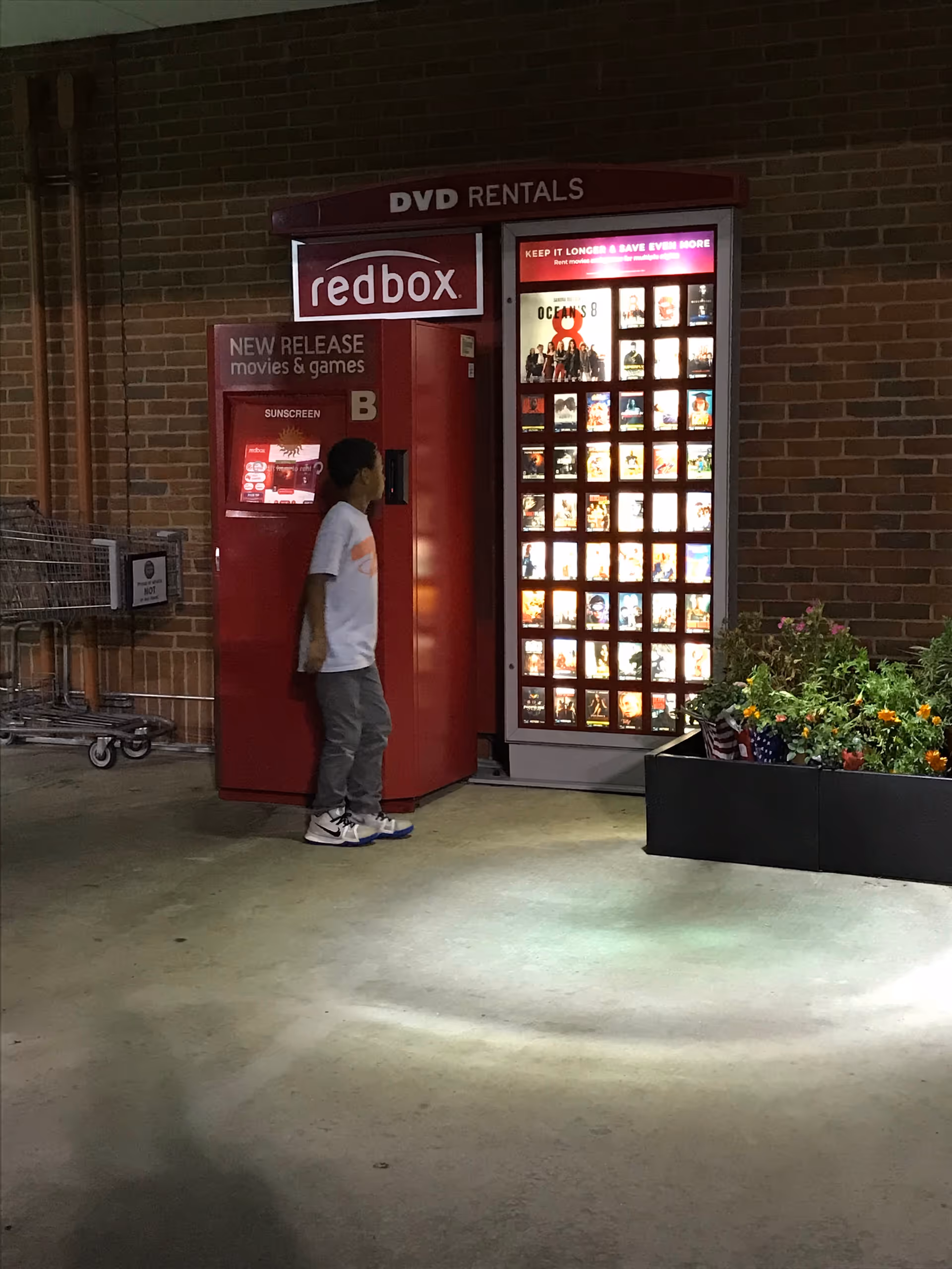 A person stands beside a redbox DVD vending machine outside a brick building.