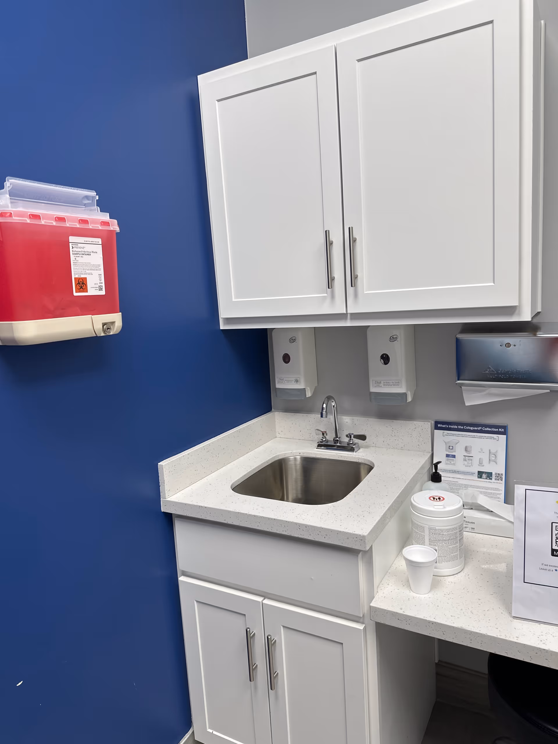 A small stainless steel sink with white cabinets on a blue wall in a clinical kitchenette.