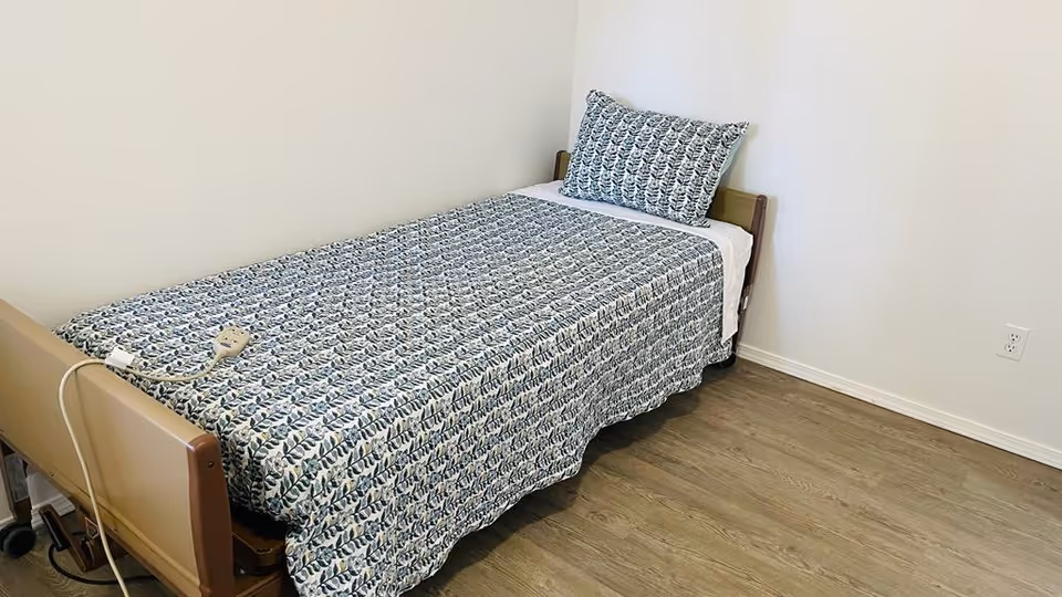 Single bed with blue patterned bedding in a small, sparsely furnished senior care room.