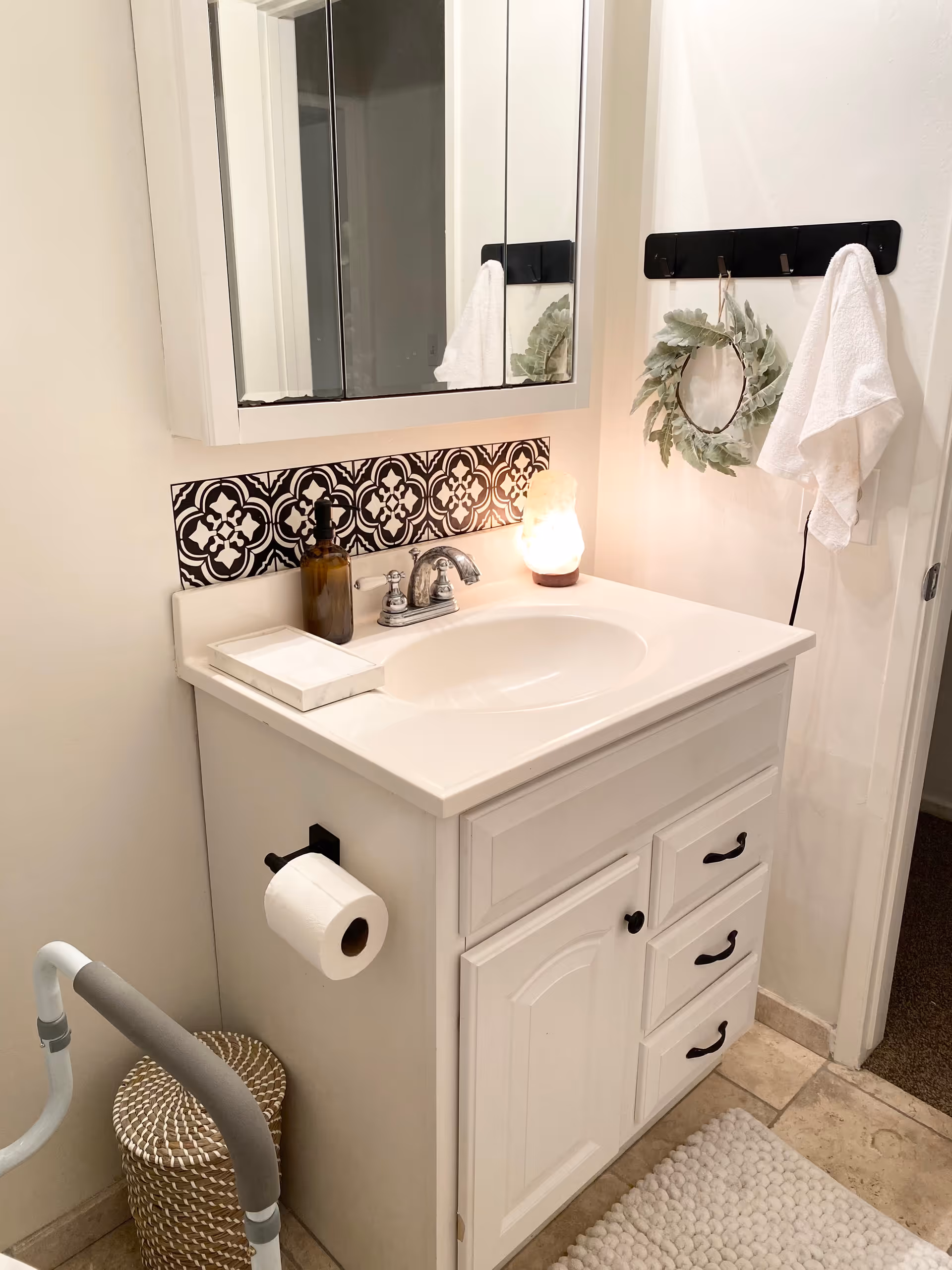 White bathroom vanity with sink, mirror, towel rack, wreath, and lit patterned backsplash.