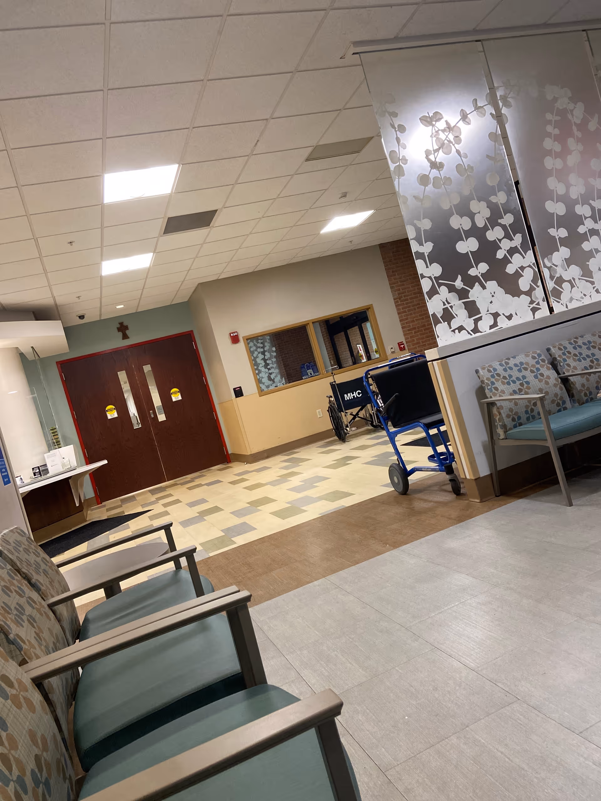 Hospital interior with tiled floors, waiting chairs, and a wheelchair near decorative panels.
