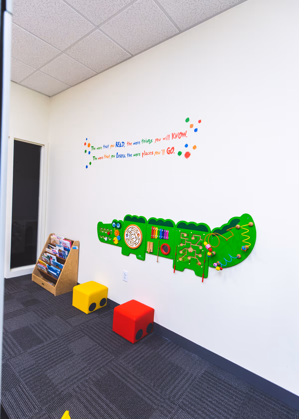 Interior play area with a green wall activity panel, magazine rack, and colorful stools.
