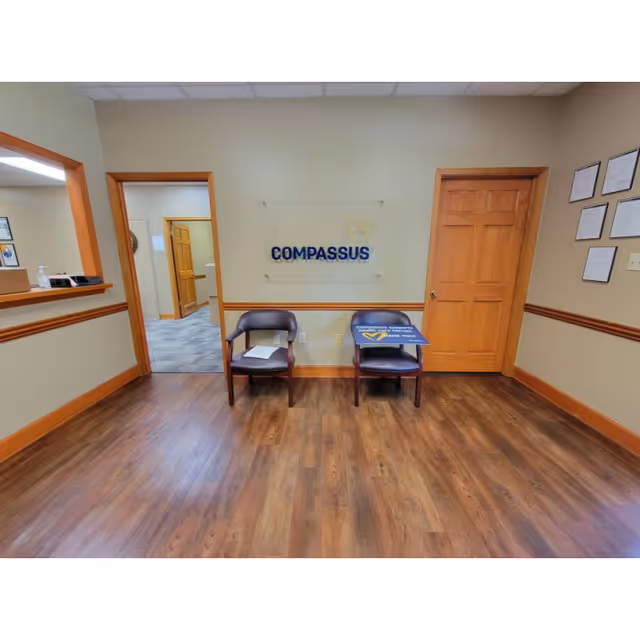 Wood-floored lobby with two chairs and COMPASSUS sign on wall.