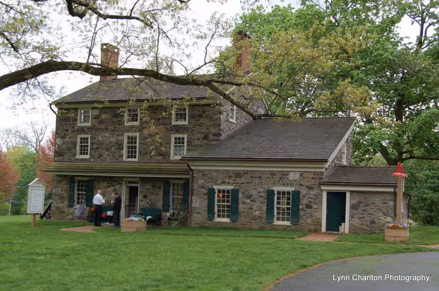 Stone building with green shutters on a grassy lawn and people at the entrance.
