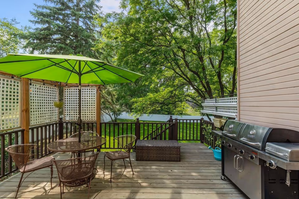 Outdoor wooden deck with green umbrella, metal table and chairs, grill, and lush trees.